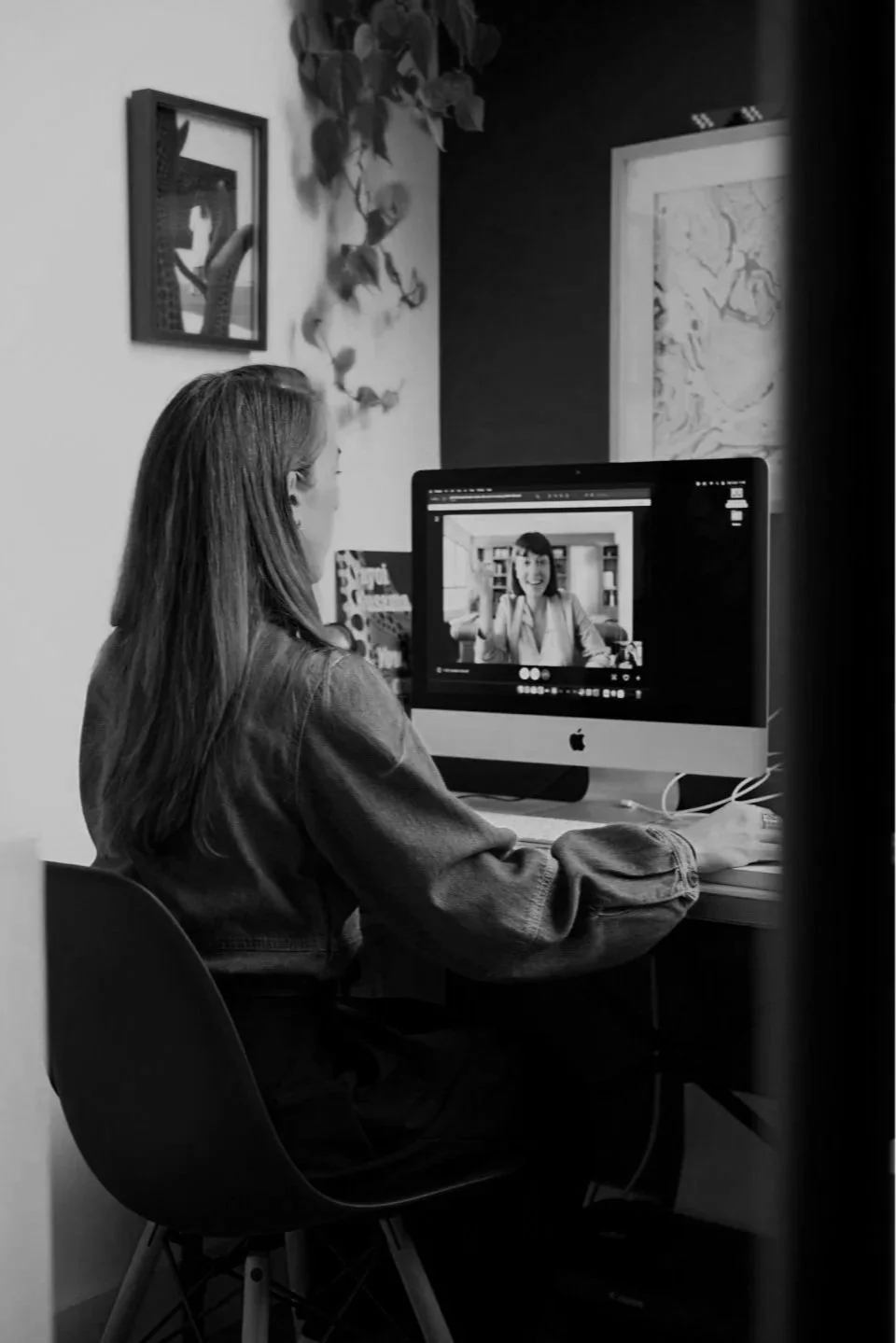 A woman with long hair sitting at a desk, working on an iMac computer. The screen shows her video call with a woman smiling and gesturing. The room has framed pictures and a plant on the wall.