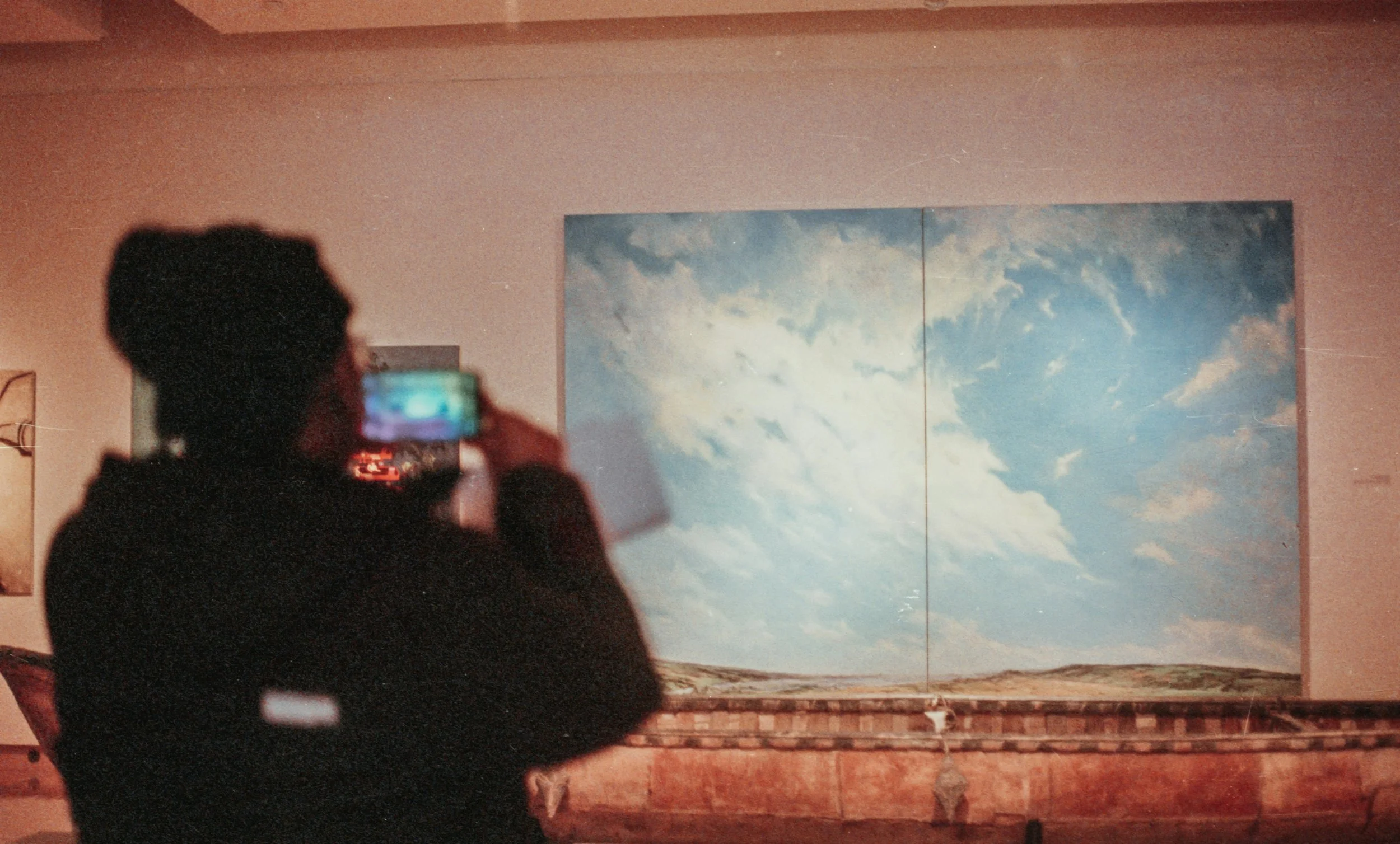 Person taking a photo of a sky painting on a wall in an indoor setting.