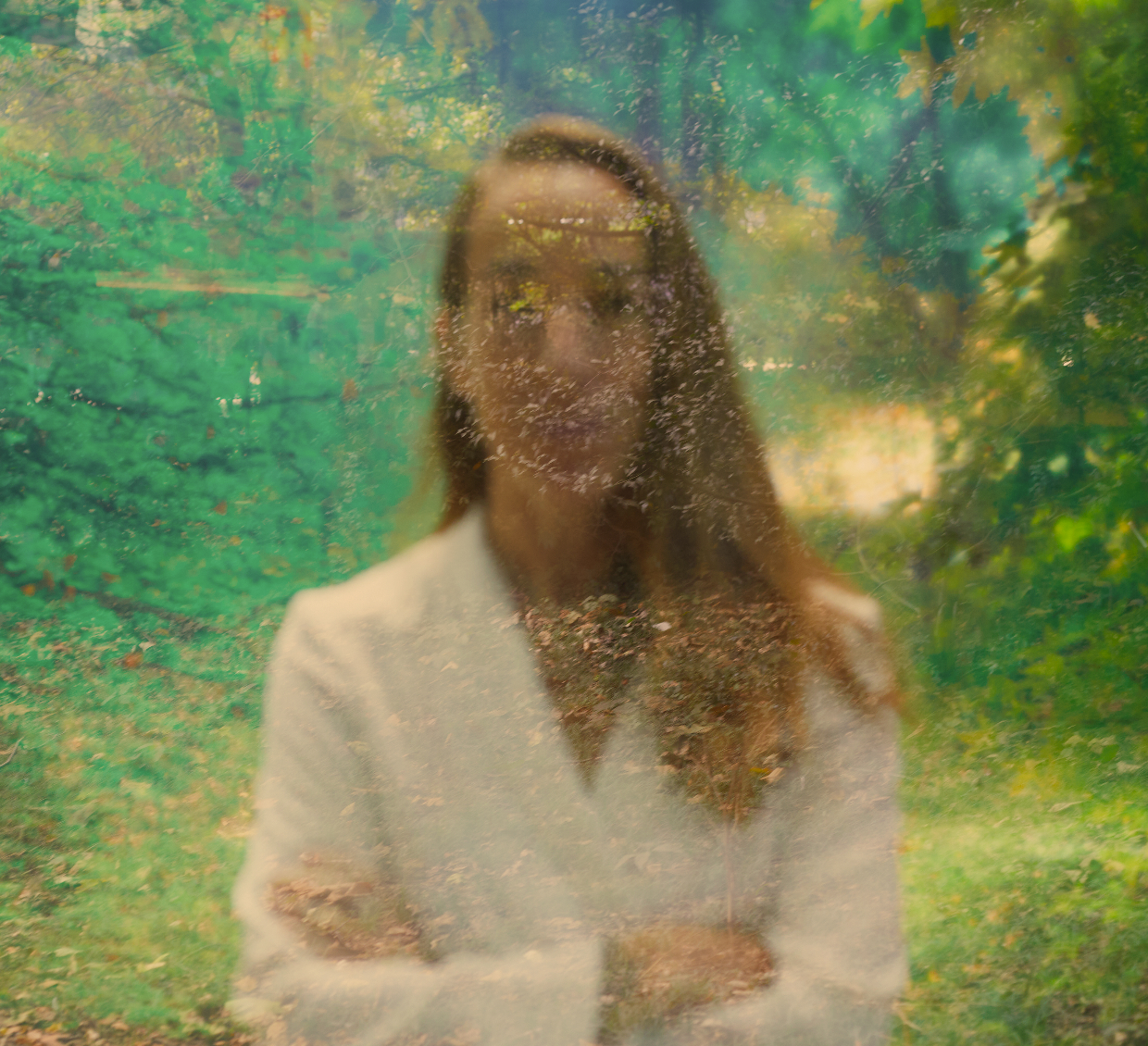 A woman with long hair wearing a white blazer is seen through a window with water droplets on it, in a green outdoor setting.