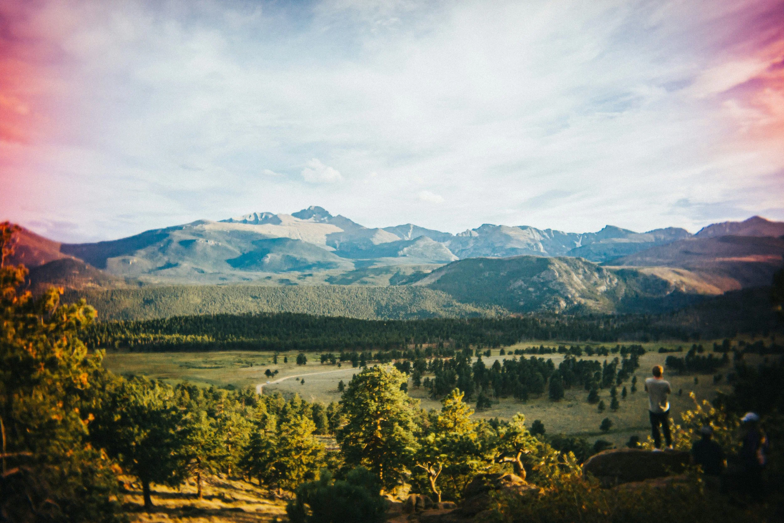 A landscape scene with distant mountains under a cloudy sky, lush green fields, and trees in the foreground. A person is sitting on a rock on the right side, facing away and looking towards the mountains.