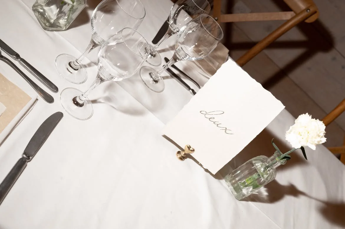 Une table élégante avec des verres à vin, une carte de place et une petite vase avec une fleur blanche sur une nappe blanche.