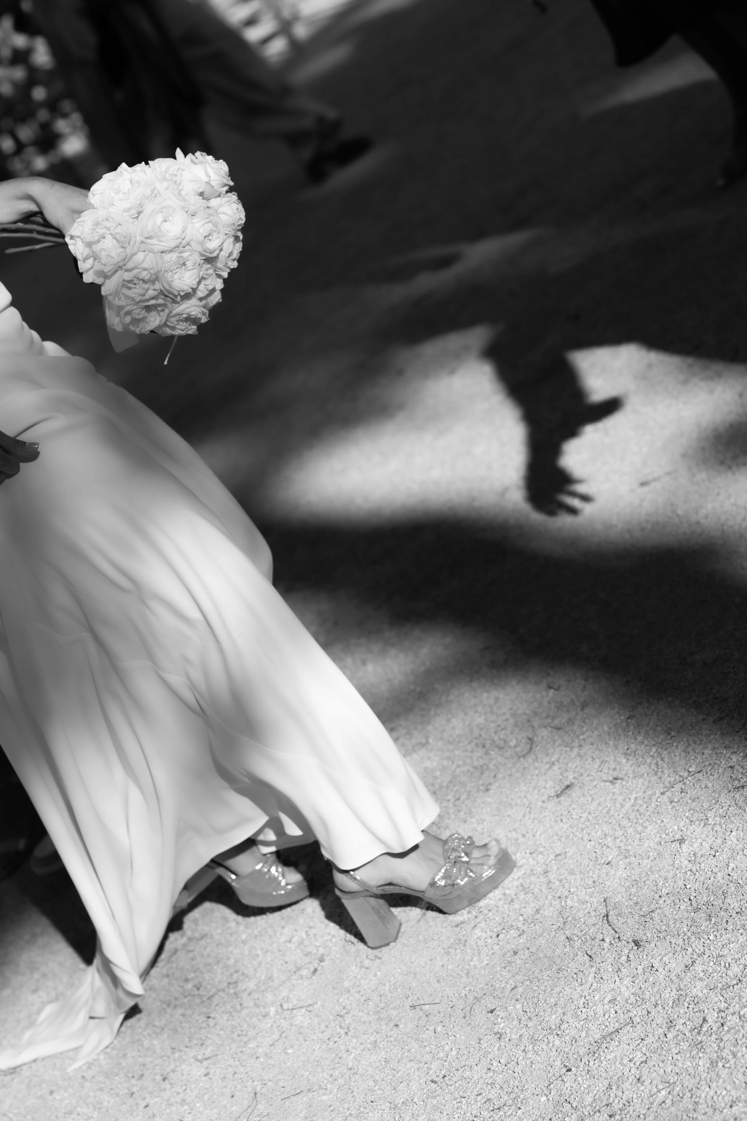 Une personne habillée en blanc, portant des chaussures à talons, assise par terre sur une surface en béton, tenant un bouquet de fleurs blanches. La photo est en noir et blanc et la personne porte une robe longue ou un vêtement fluide.
