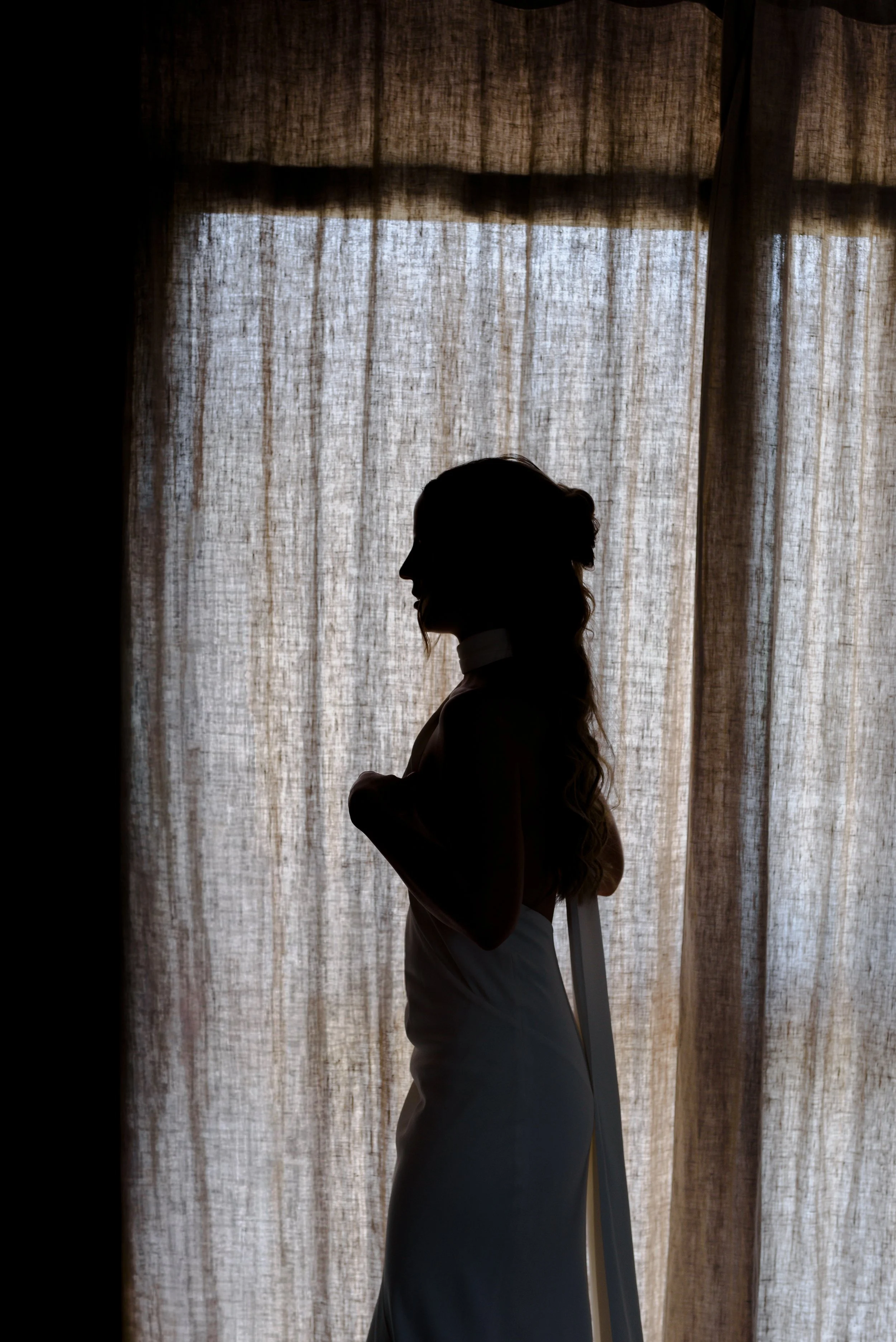 Silhouette d'une femme enceinte debout près d'une fenêtre avec des rideaux en tissu léger, au coucher ou lever du soleil.