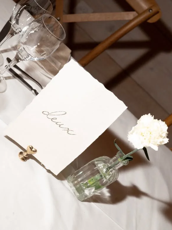 Une table élégante avec une bouteille en verre contenant une fleur blanche, un menu avec le mot 'eau' écrit dessus, et des coupes à vin en arrière-plan, dans un cadre chaleureux.