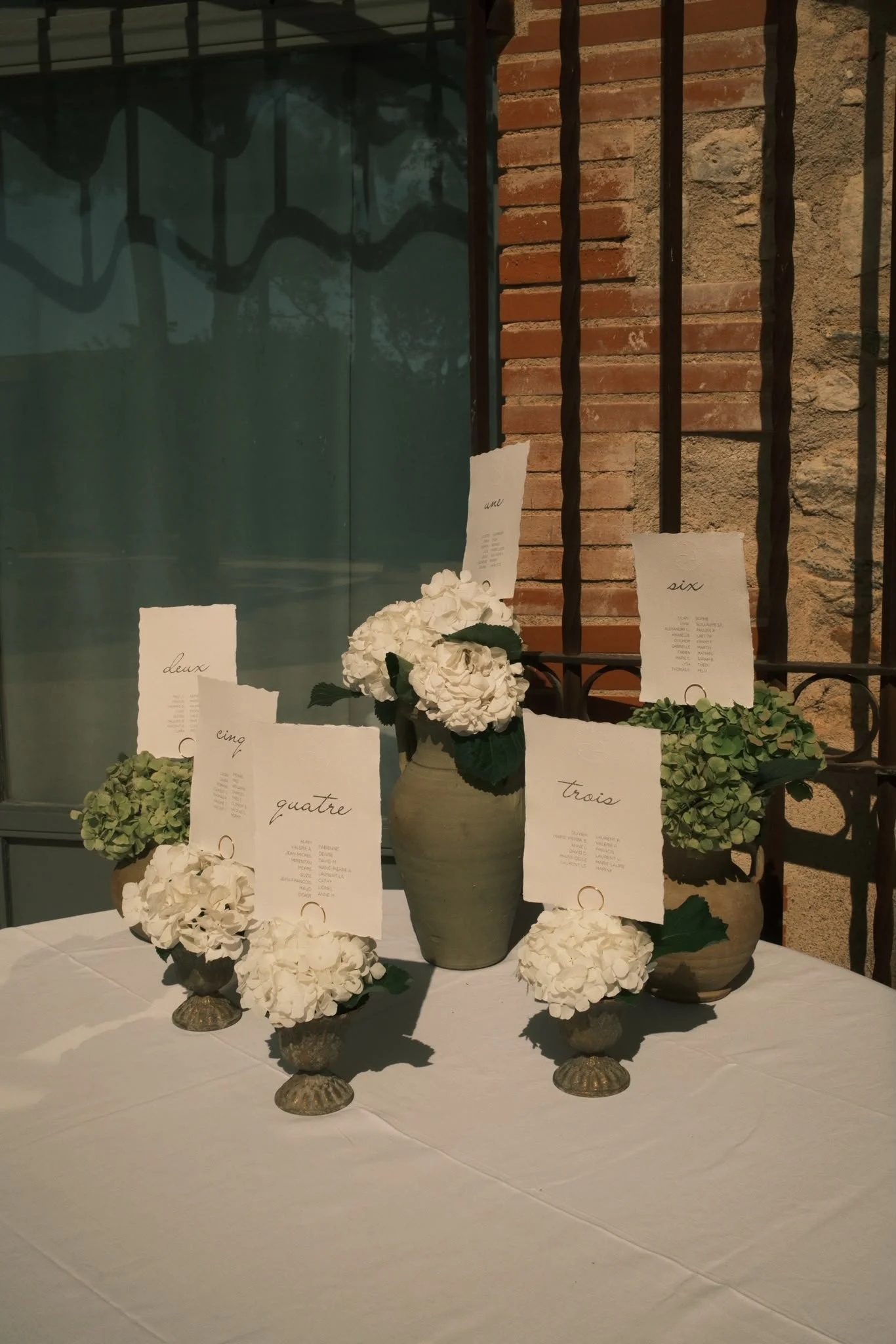 Arrangement de vases avec des bouquets de fleurs blanches, entourés de papiers avec des numéros écrits en français, sur une table blanche. Le décor en arrière-plan comprend un mur en briques et des barres en fer.