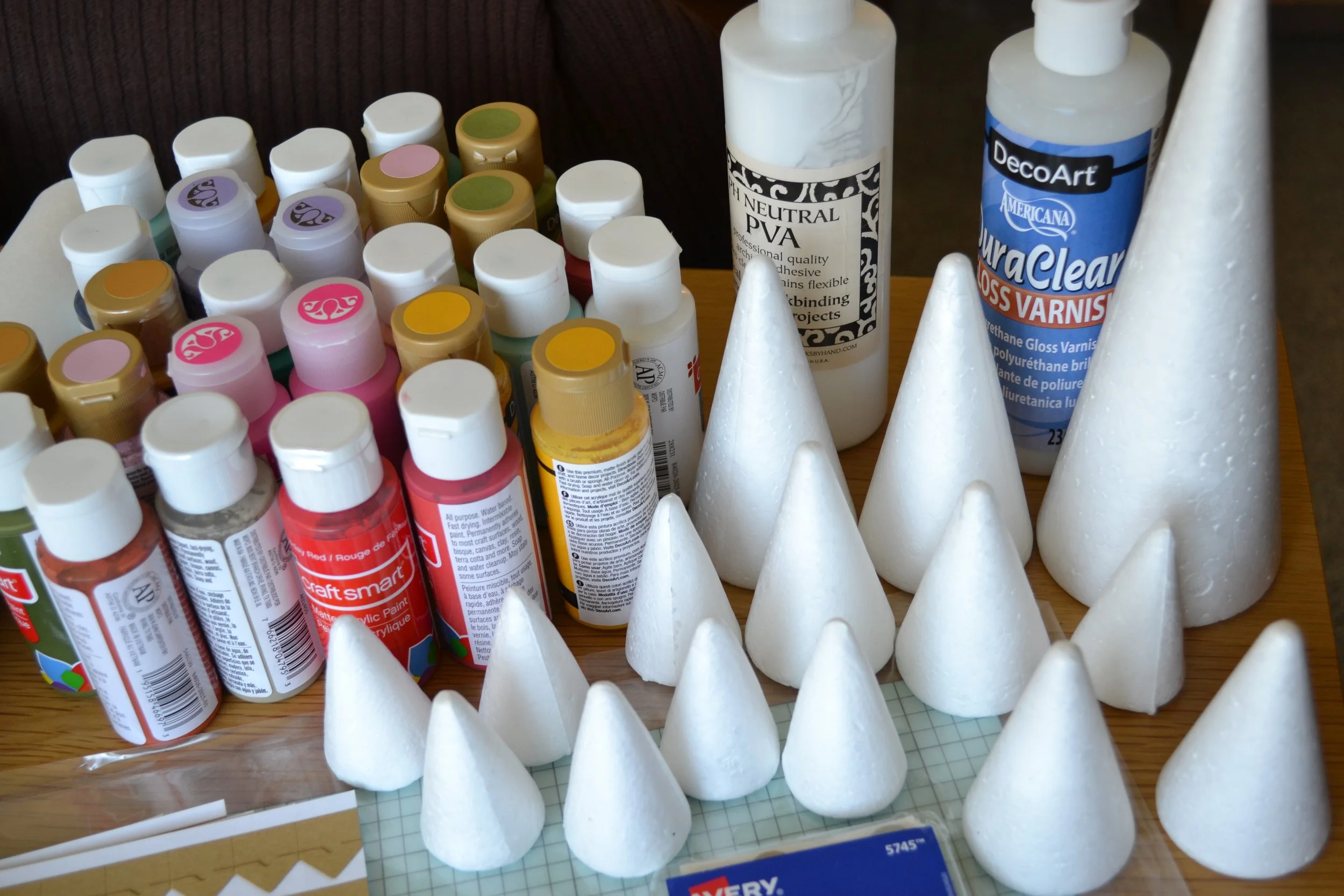 Assorted bottles of acrylic paint, glue, and spray varnish on a wooden surface, with white foam cone shapes in the foreground, possibly for craft or art project preparation.
