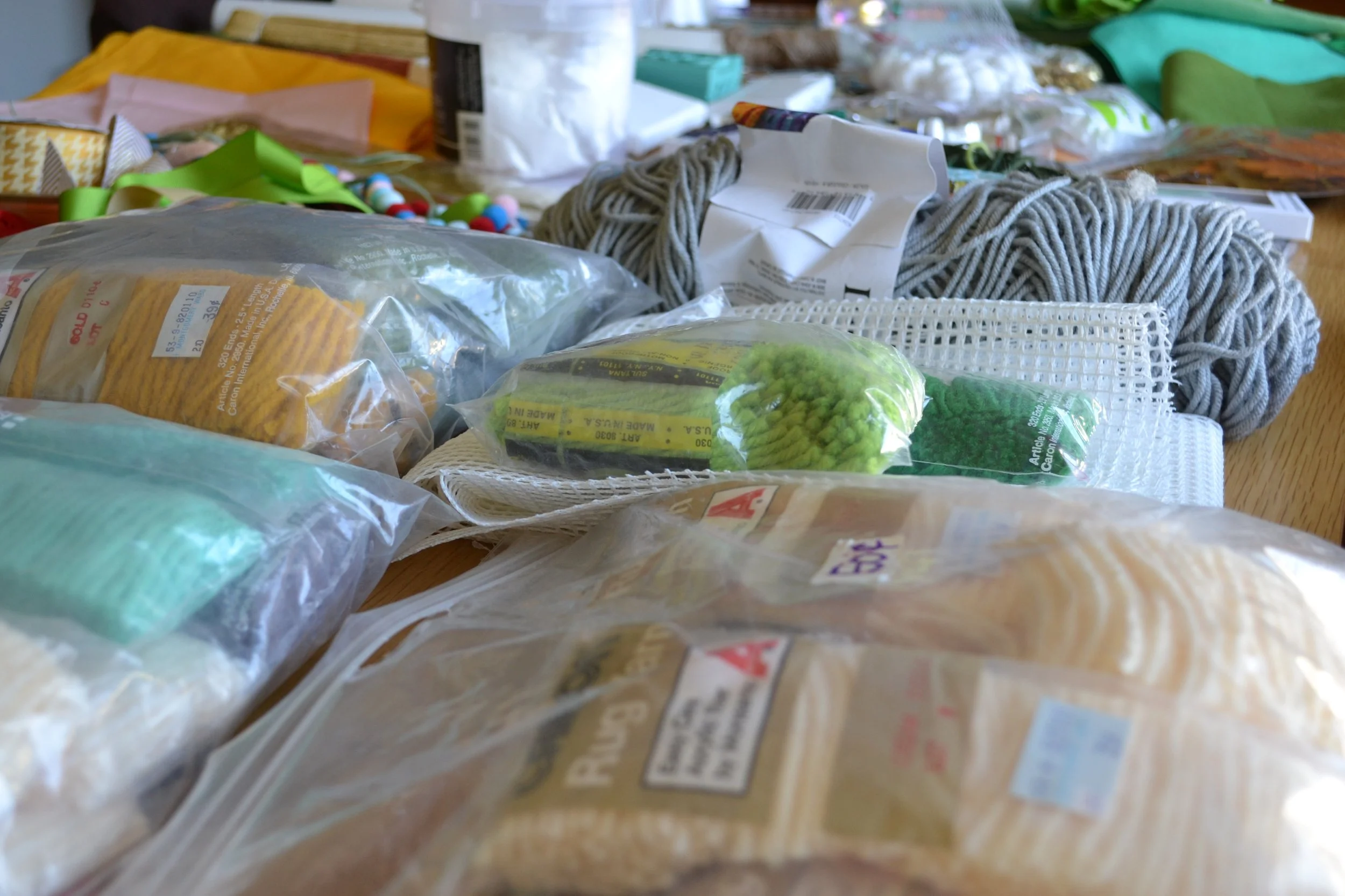 Cluttered table with various craft supplies including yarn, foam pieces, plastic bags, and packaging materials.