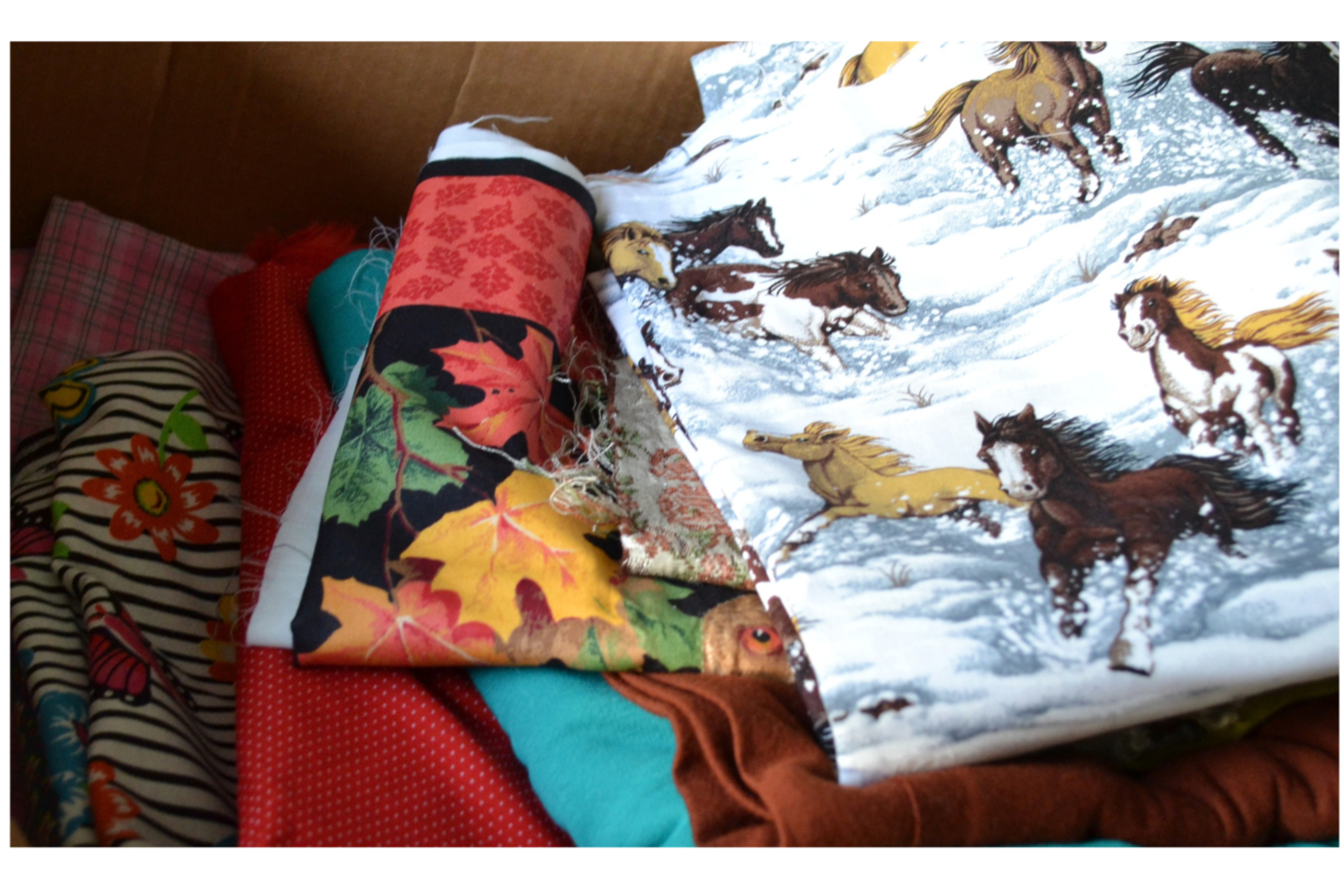 Stacked fabric pieces with various patterns, including horse and winter scene designs, on a bed with a pink and brown bedding.