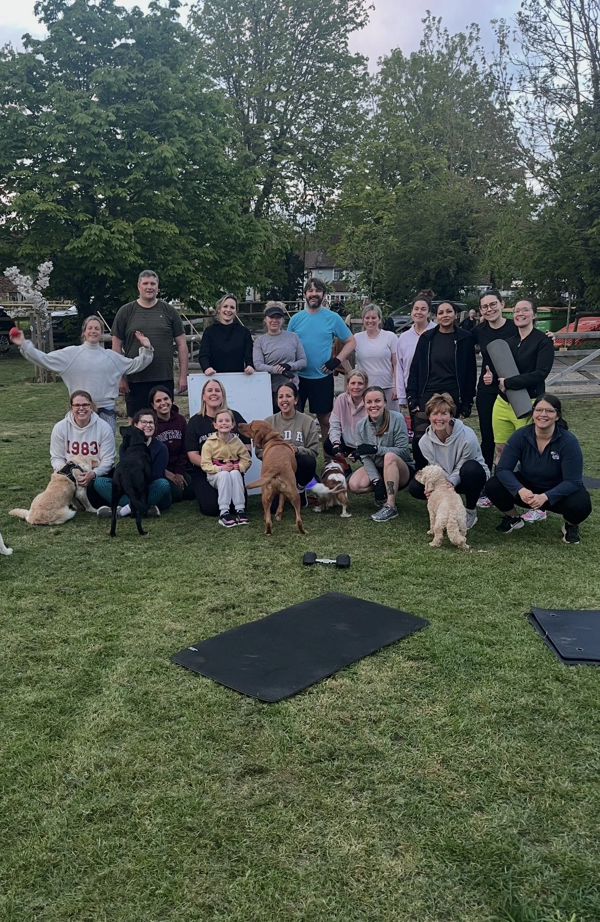 Group of people and dogs gathered outdoors on a grassy field, with trees and a fence in the background, some holding equipment and a whiteboard.