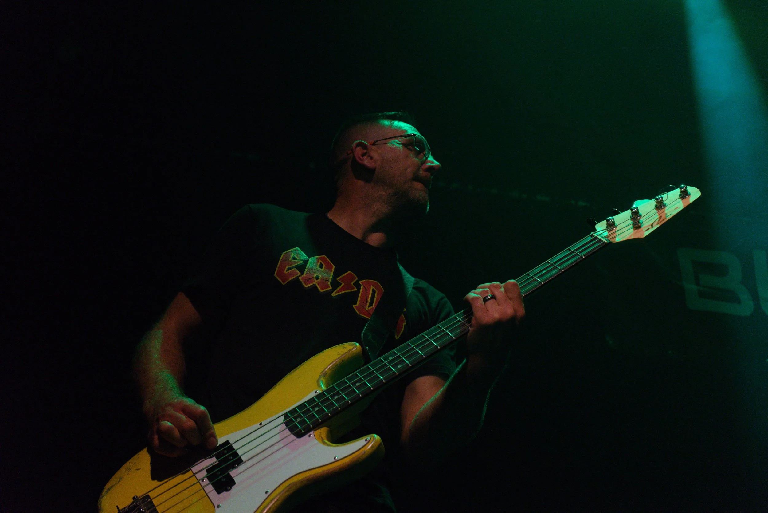 A man with glasses and a beard playing an electric bass guitar on stage, illuminated by green stage lighting, wearing a black T-shirt with red, yellow, and white lettering.