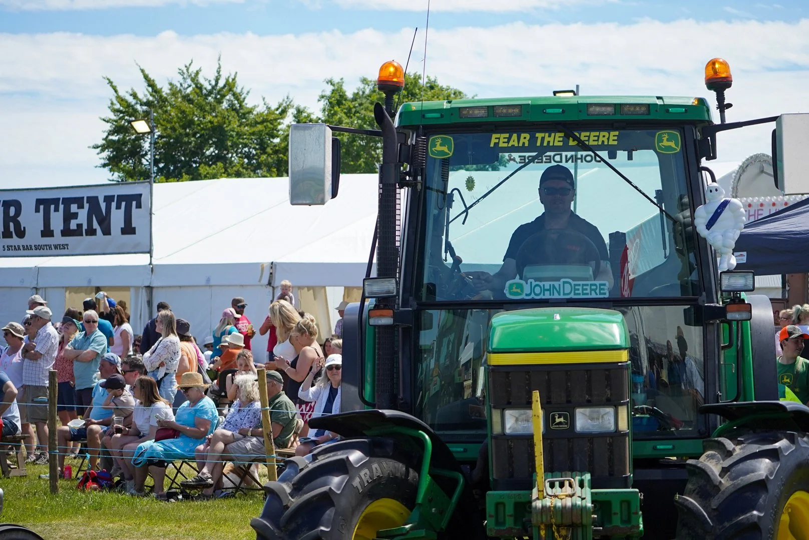 A large John Deere tractor at an outdoor fair or festival with a crowd of people sitting and standing nearby, some under white tents. The tractor has a sign with yellow text saying "FEAR THE REEE" and an inflatable Michelin Man attached to the right 
