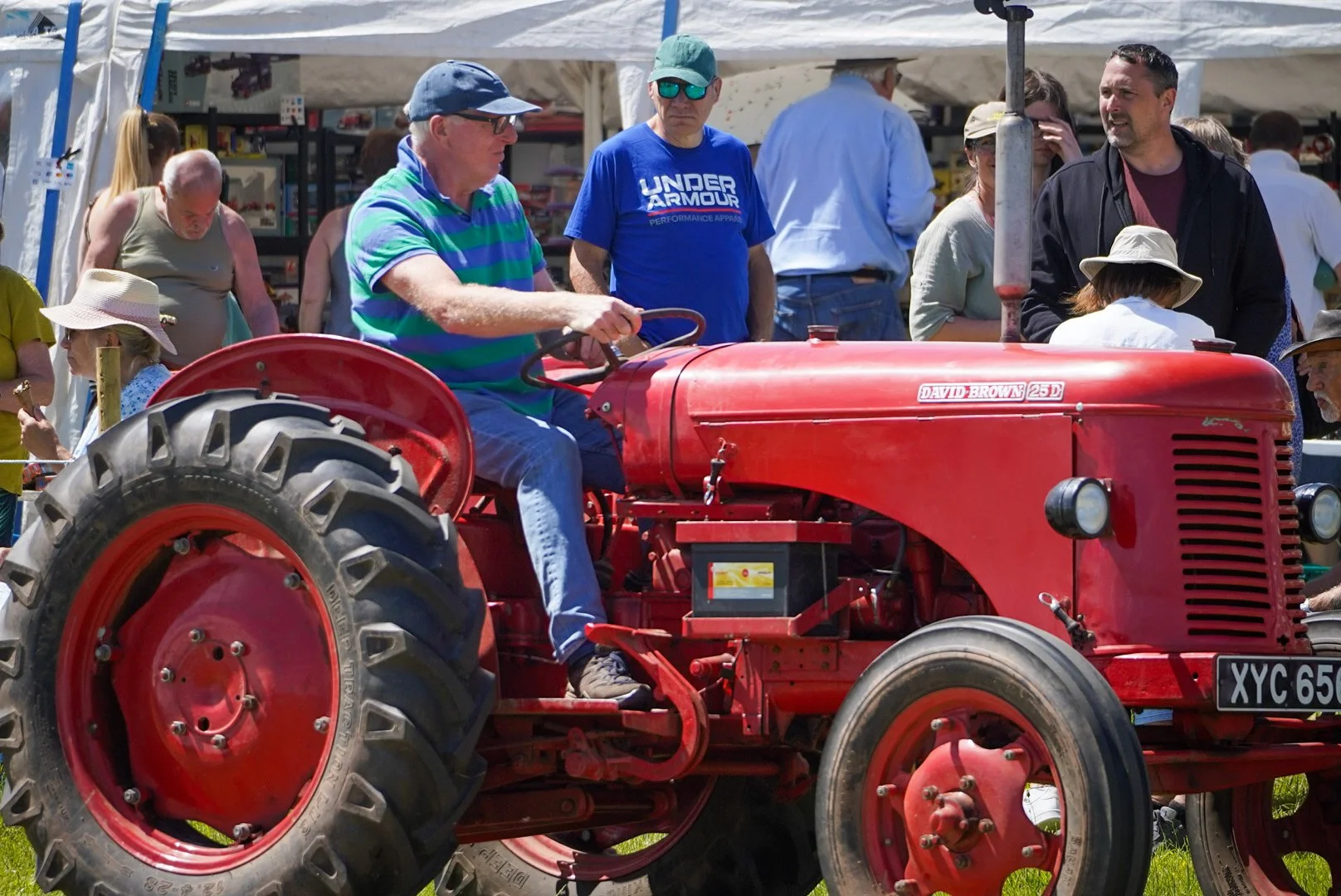 A man is sitting on a red vintage tractor, surrounded by a crowd of people at an outdoor event. The man is wearing a striped shirt, jeans, and a cap, and is holding the steering wheel of the tractor. The crowd includes women and men, some wearing hat