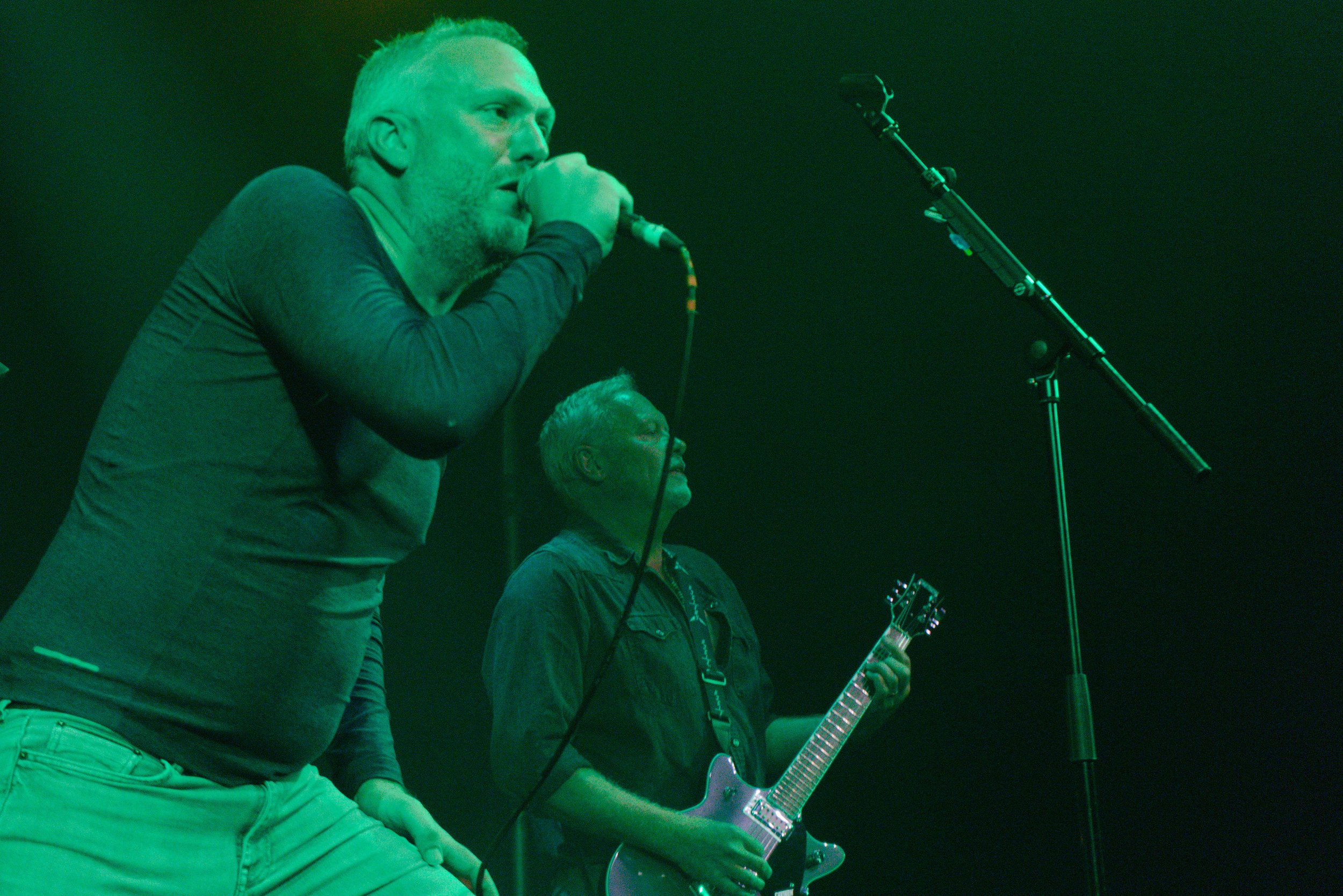Two men on stage performing, one singing into a microphone and the other playing an electric guitar. The scene is lit with green stage lighting.
