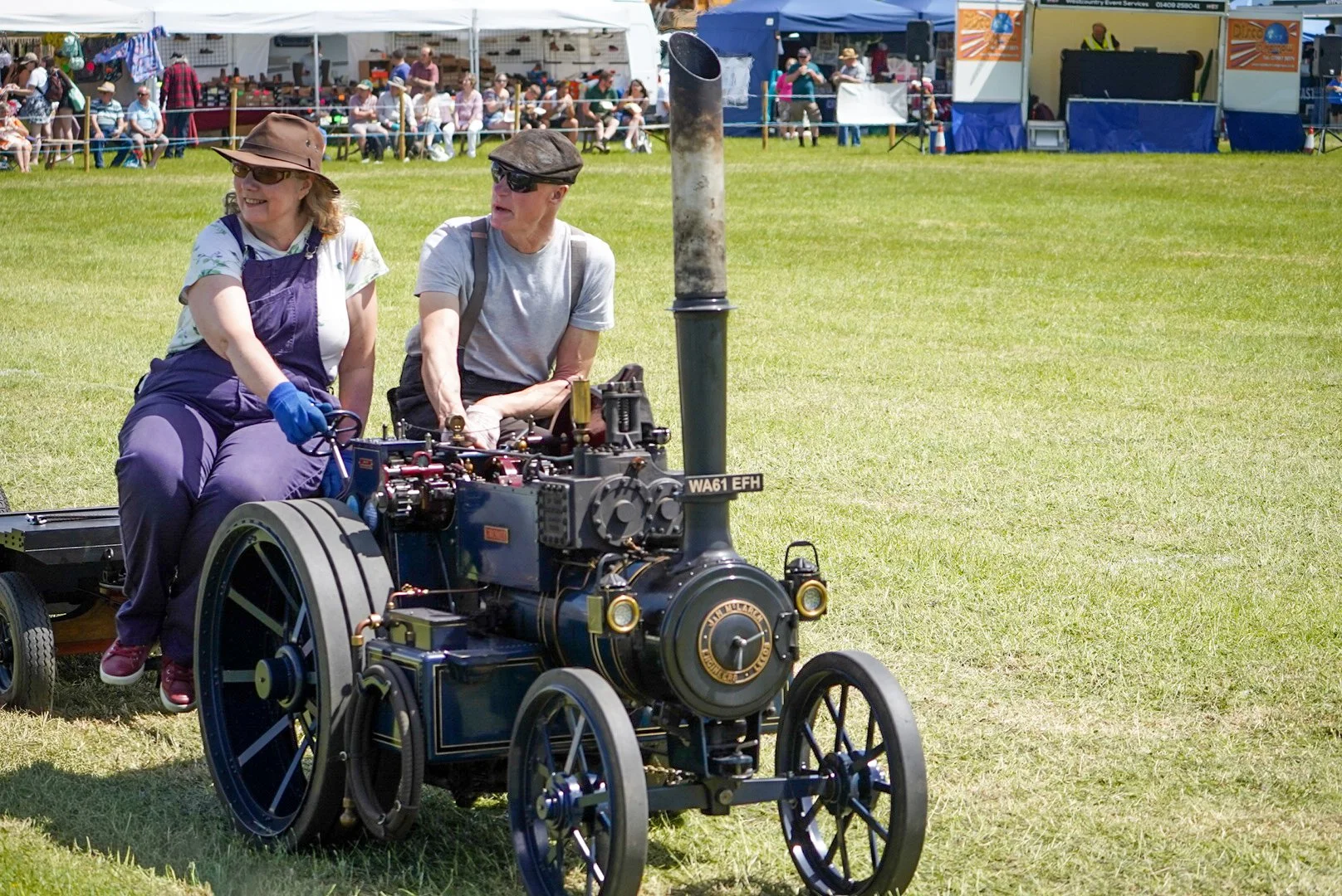 Two people sitting on a vintage steam engine at an outdoor event, with booths and a crowd in the background.