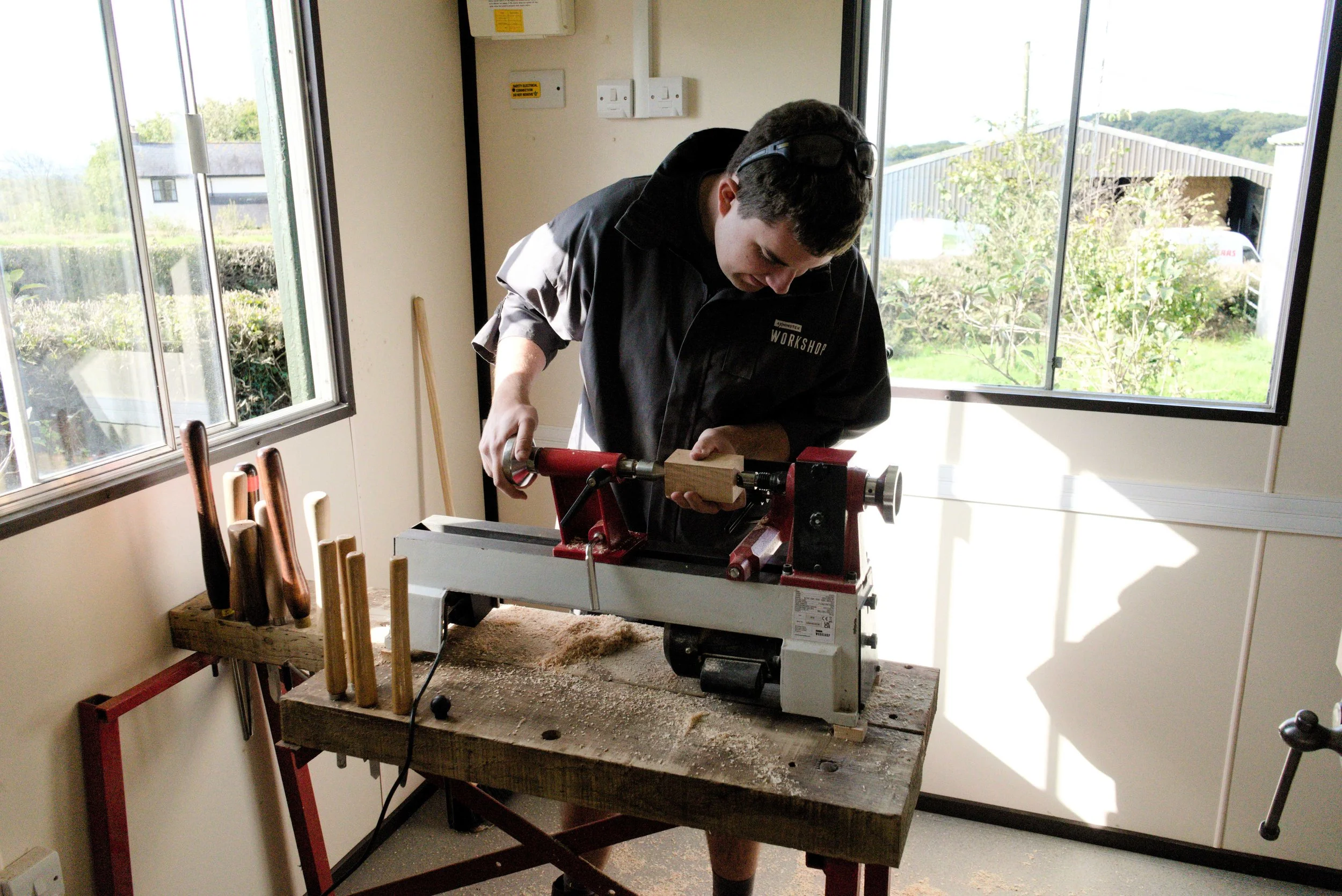 A young man in black protective glasses and a dark workshop shirt is working with a woodworking machine on a piece of wood in a bright workshop with large windows showing greenery outside.