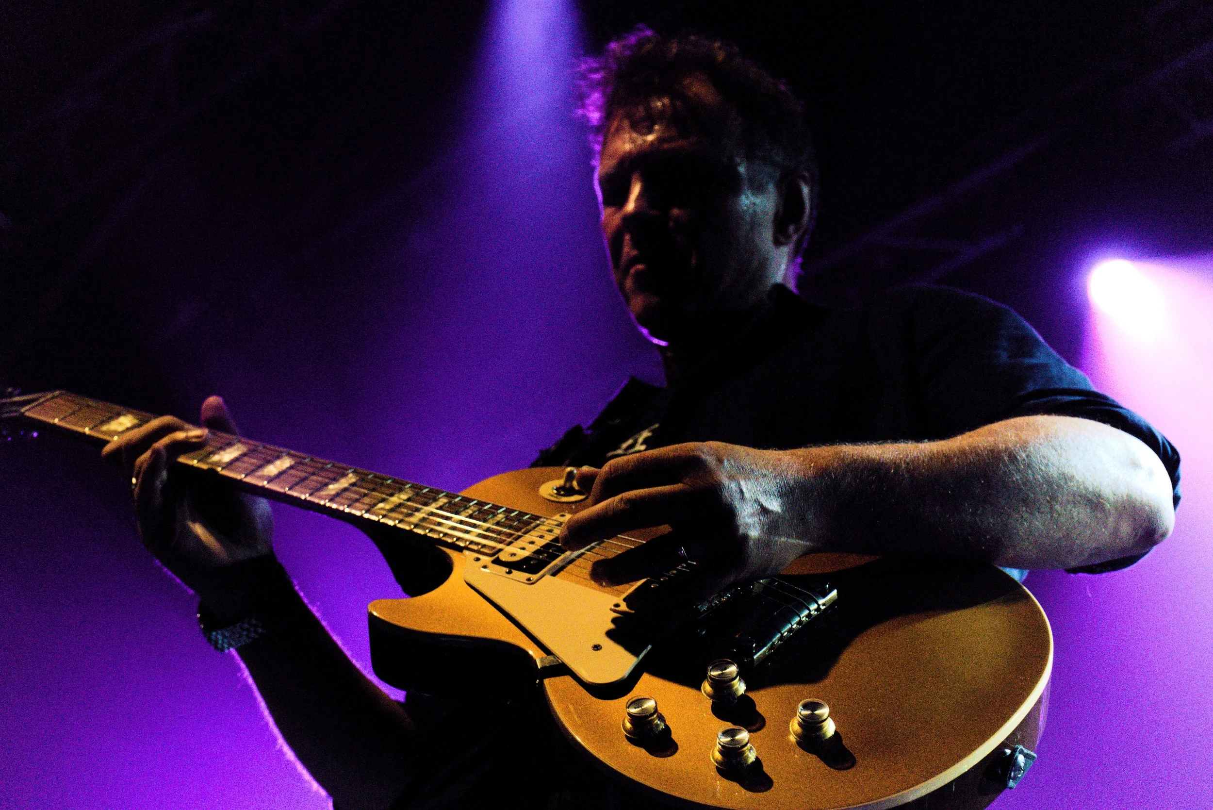 A man playing an electric guitar on stage, illuminated by purple and blue stage lights.