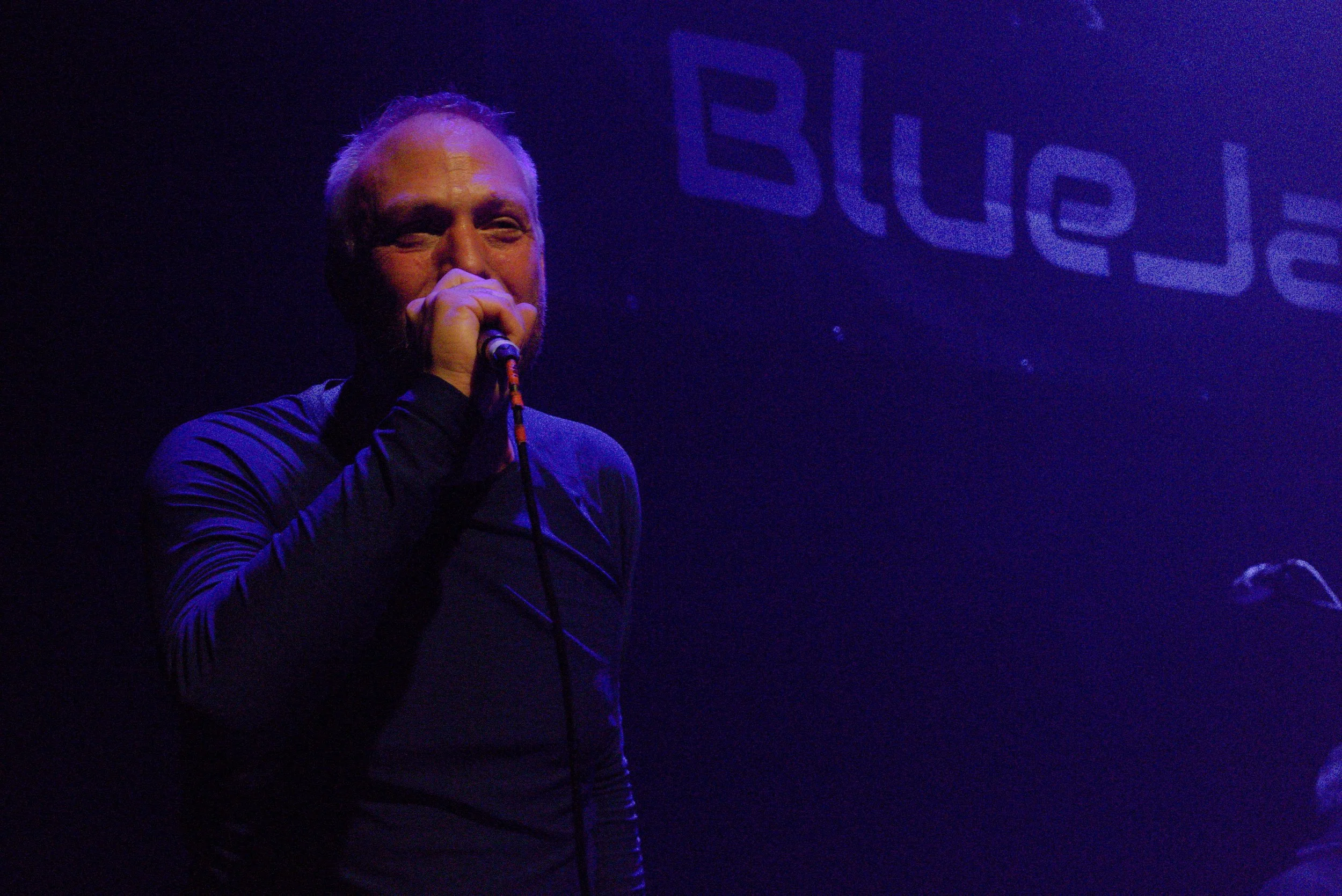 A man with gray hair and a beard singing into a microphone on stage, illuminated by purple lighting, with a large illuminated sign in the background that says 'BLUE'.