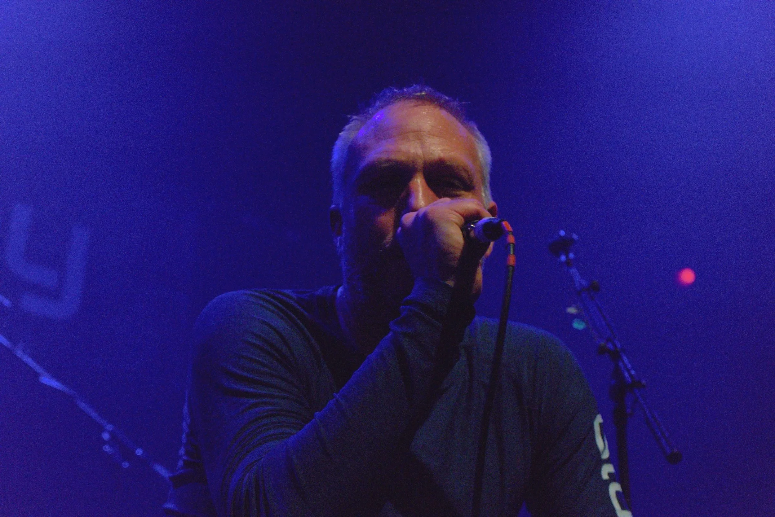 Male singer performing on stage with blue lighting, holding a microphone close to his mouth, wearing a dark long-sleeve shirt.