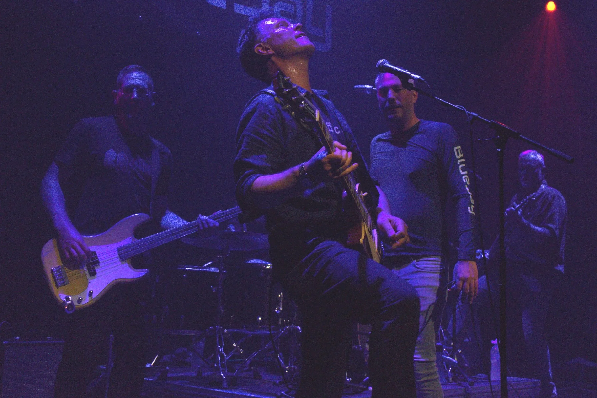 Musician on stage with a guitar, performing with three other band members under purple and red stage lighting, surrounded by musical equipment.