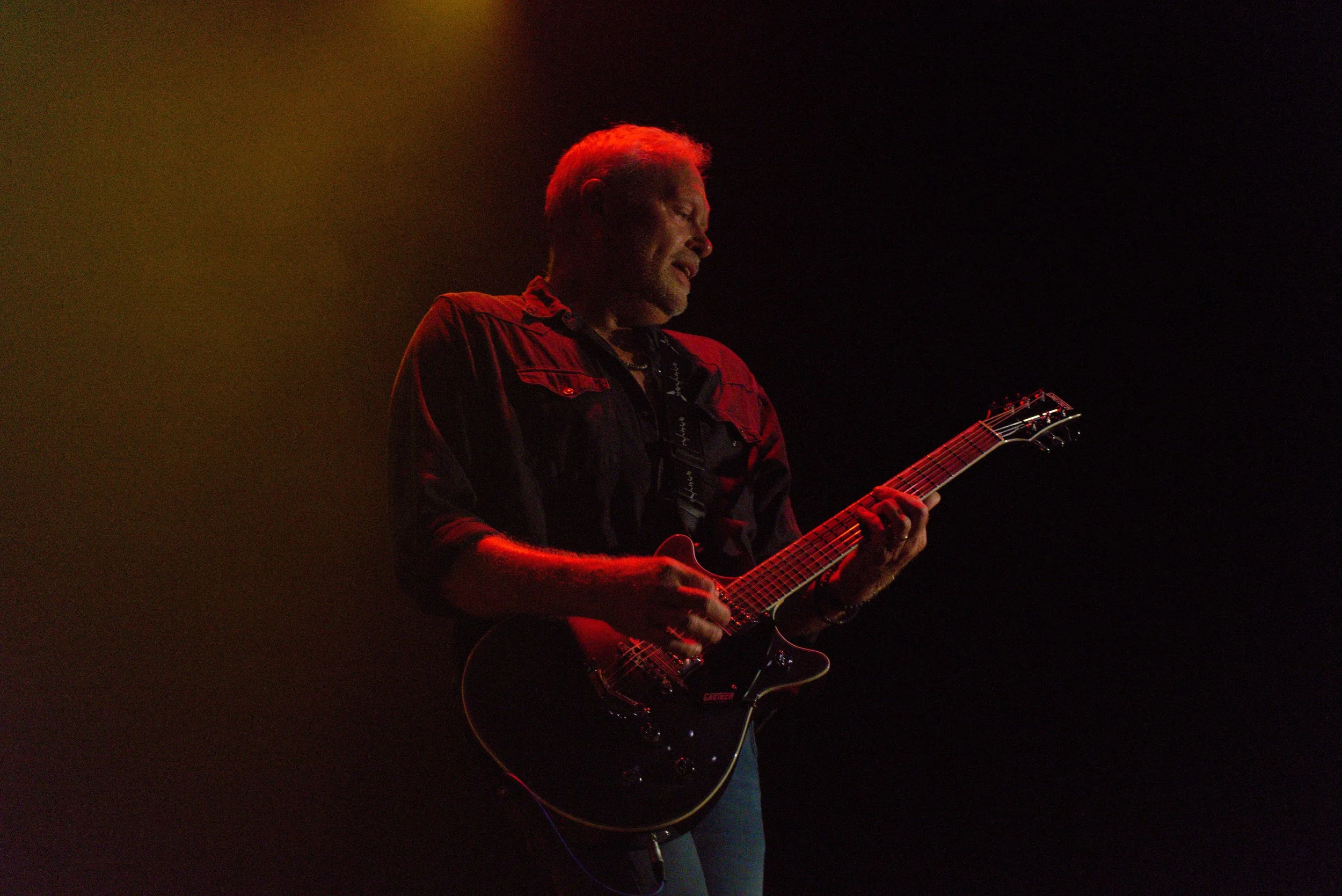 A man playing an electric guitar on stage, illuminated by red and orange lighting with a dark background.