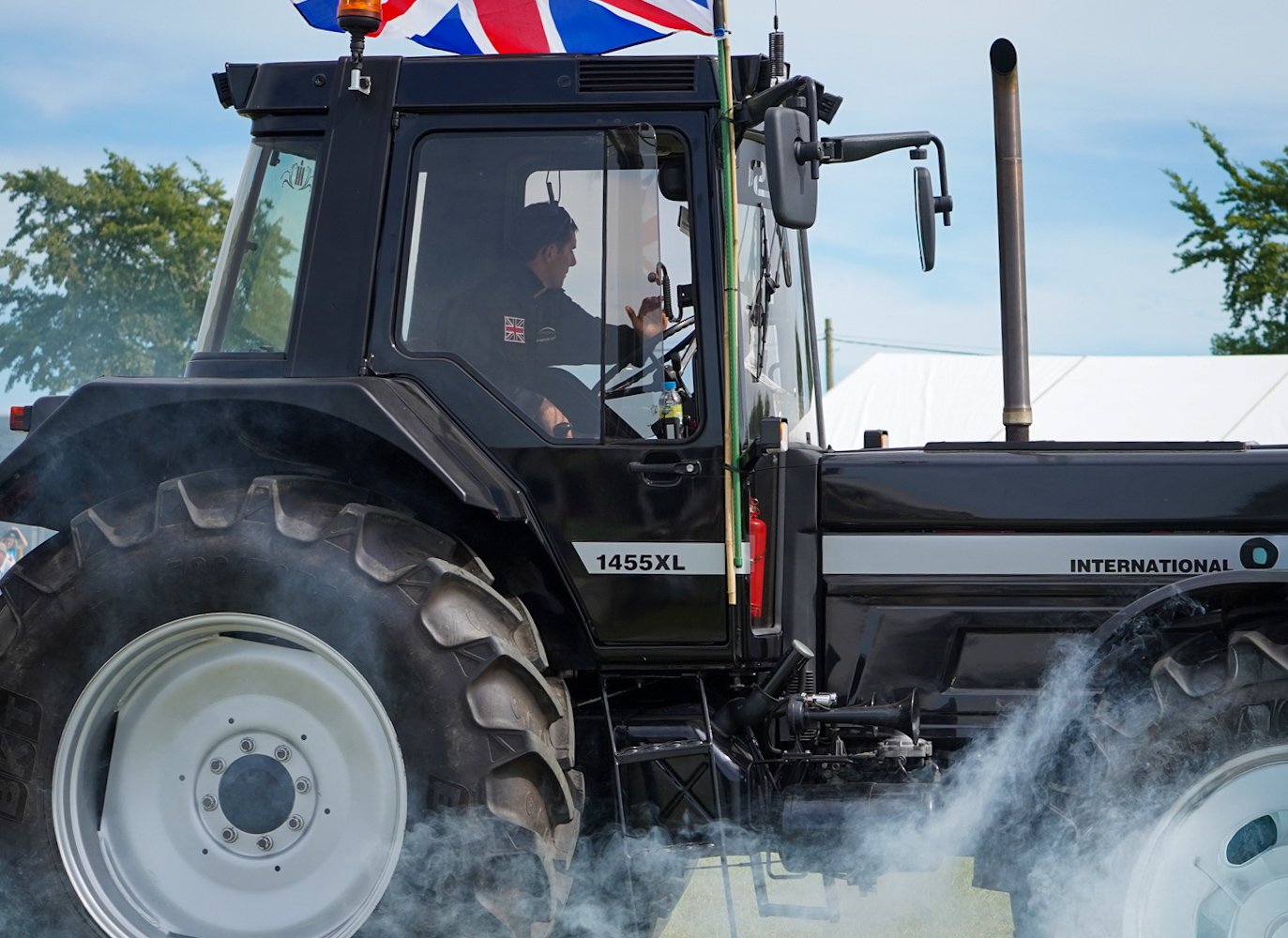 A black tractor with a British flag attached to the top, driving on a field with steam or smoke around its tires, and a person inside the cab waving.