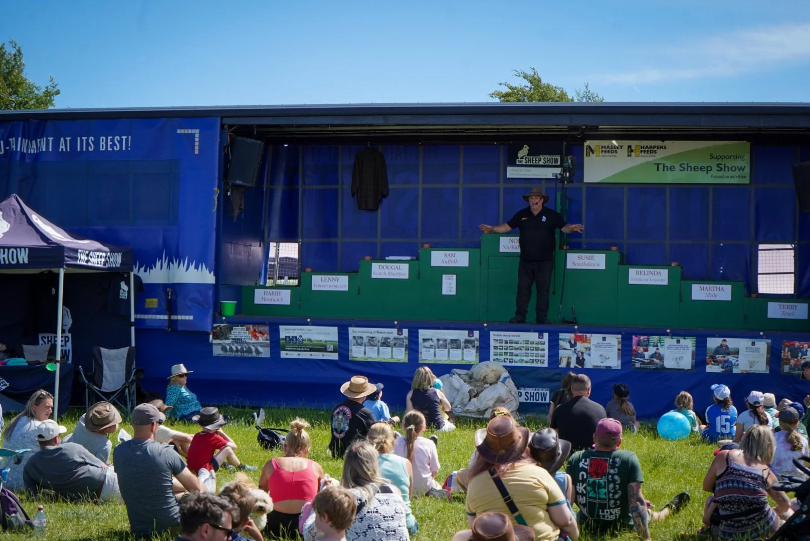 A man in black shirts and a cowboy hat is talking on a stage at The Sheep Show, with an audience sitting on the grass in front of him. The stage has multiple signs with names of sheep and sponsors, and a banner promoting The Sheep Show is visible.