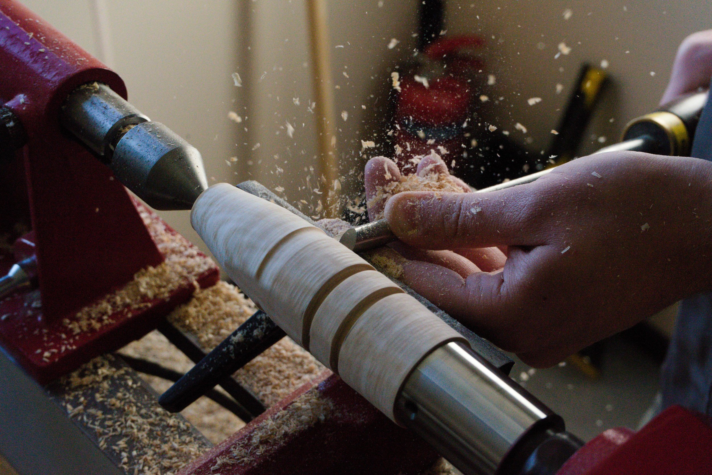 A woodworker's hands guiding a piece of wood on a lathe machine, which is shaping the wood into a round object as sawdust and wood shavings fly around.