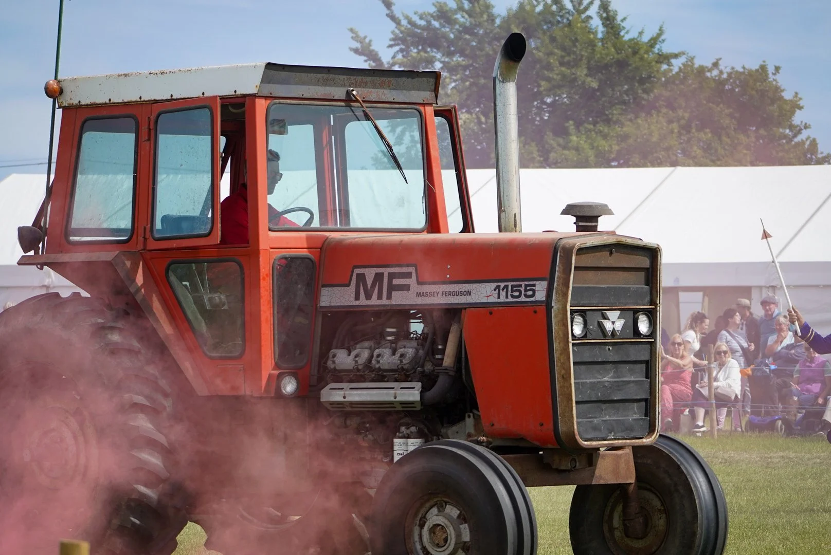 A vintage red Massey Ferguson 1155 tractor on a field, with a group of people seated under a white tent in the background during an outdoor event.