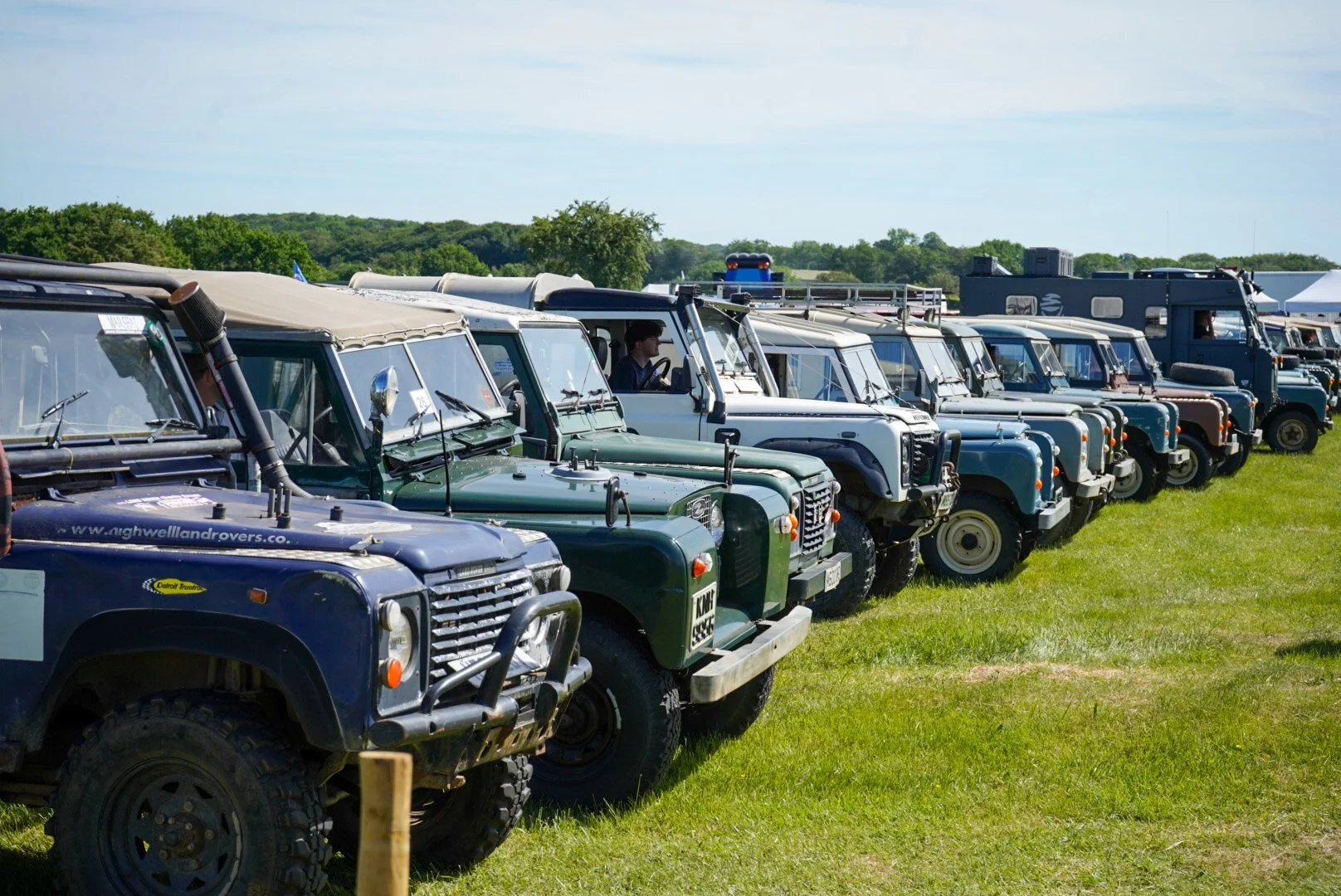 Lineup of vintage Land Rover vehicles on grass at an outdoor event under a partly cloudy sky.