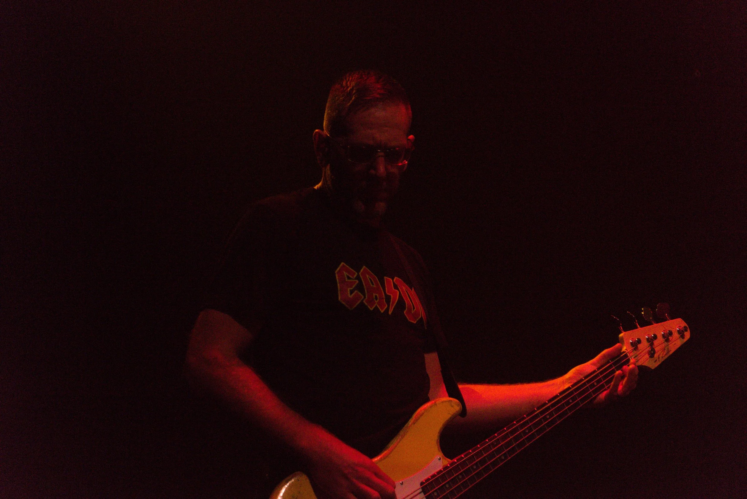 Person playing electric guitar in a dark setting with red lighting, wearing glasses and a black shirt with red and yellow text.