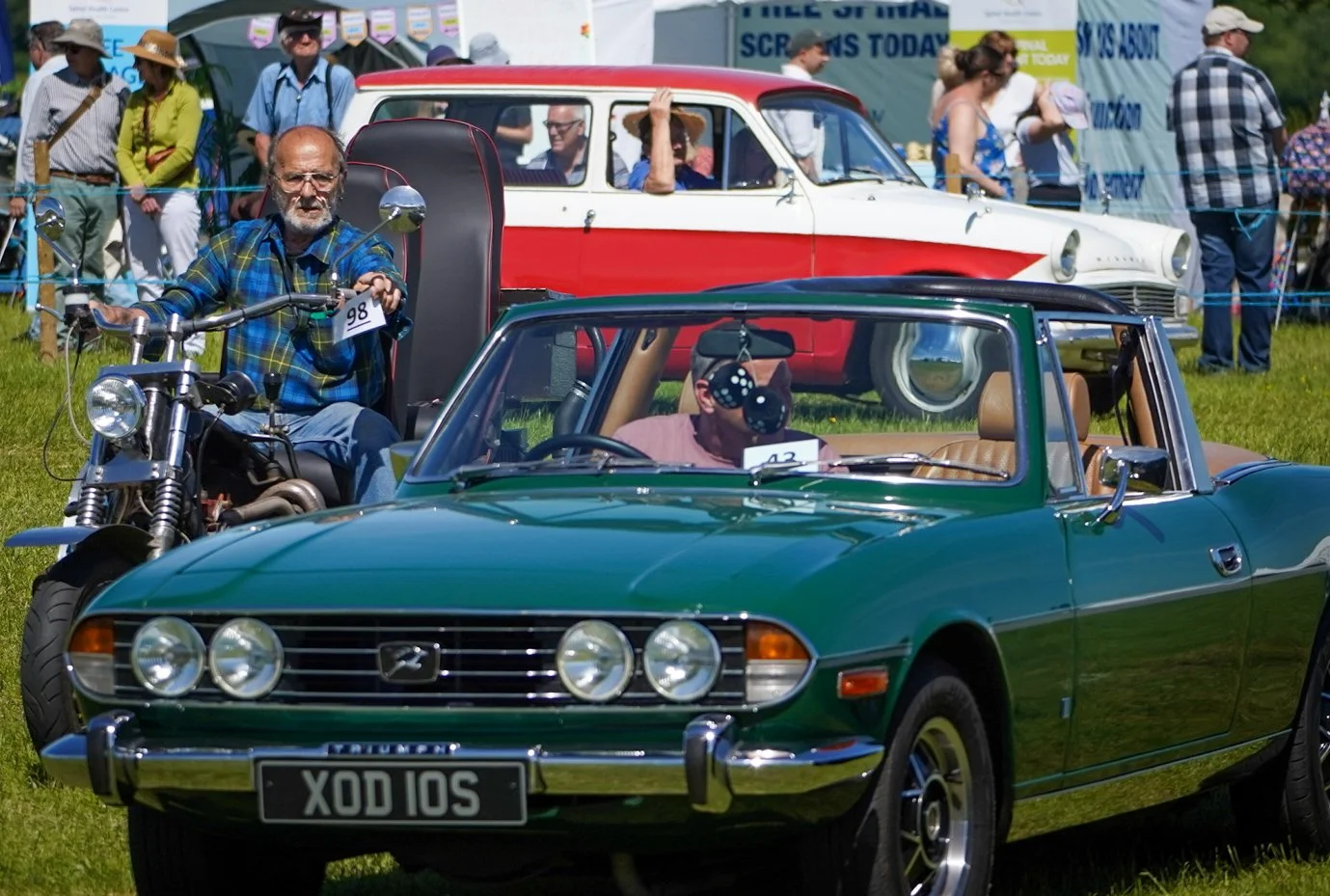 A classic green Triumph convertible car parked on a grassy field during a car show with a man driving, surrounded by people and vintage vehicles, including a red and white station wagon in the background, and a man sitting on a motorcycle with a uniq