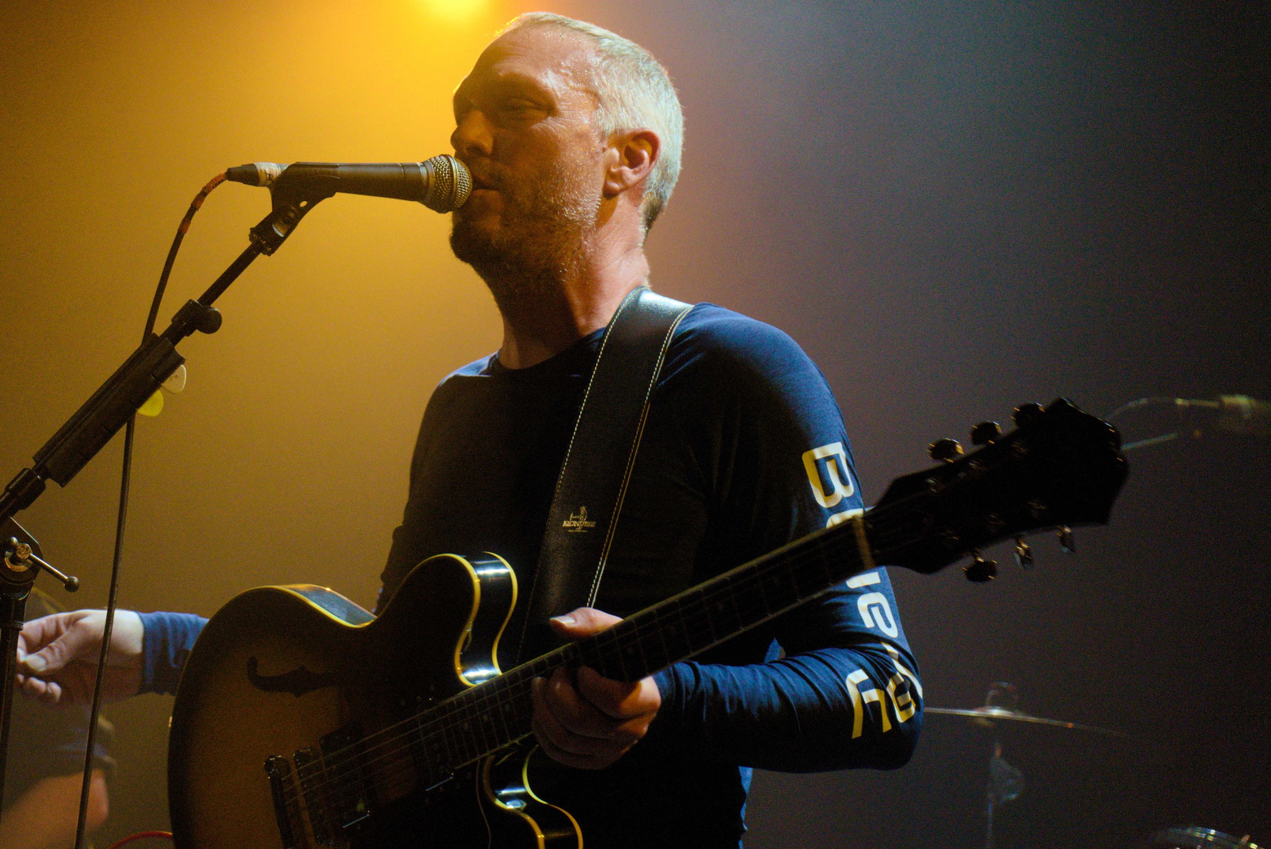 A man with short gray hair and a beard plays an acoustic guitar and sings into a microphone on stage, illuminated by warm stage lights.