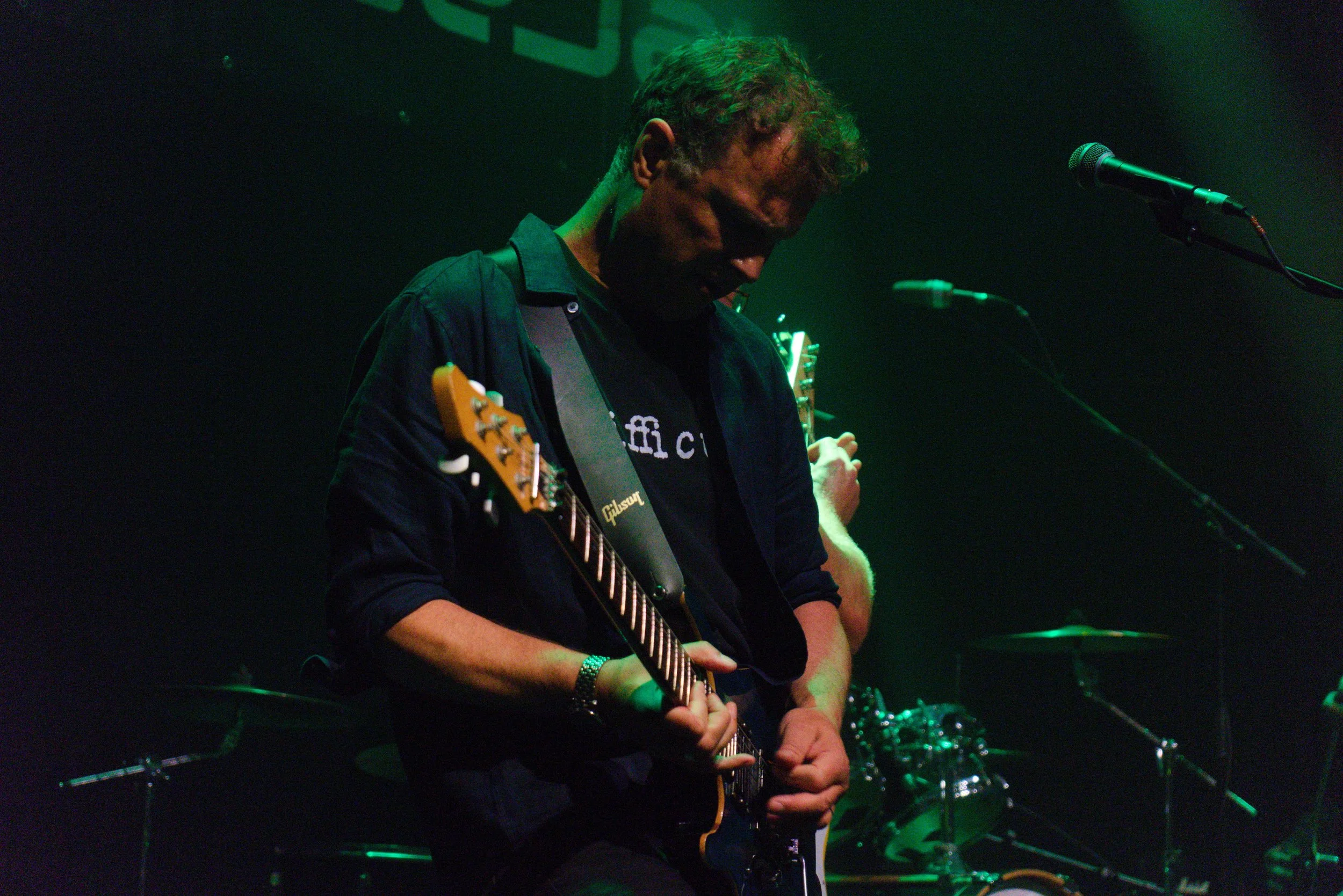 A man with curly hair playing an electric guitar on stage with green lighting, standing in front of a microphone, with a drum set visible in the background.