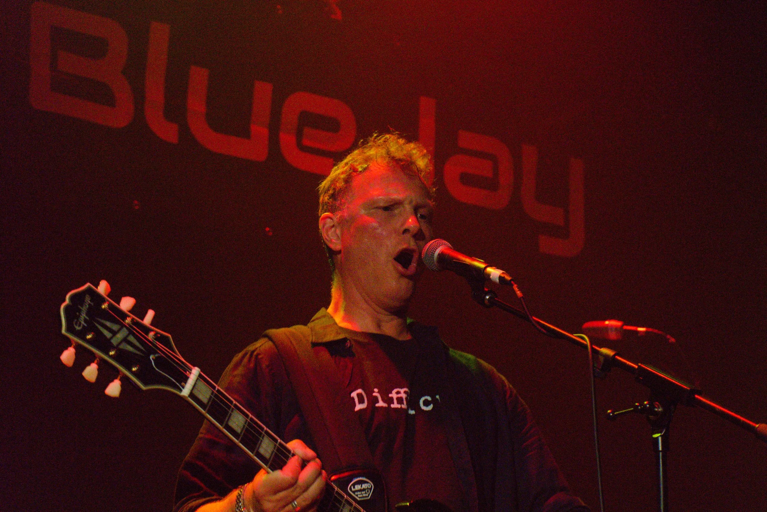 A man with light-colored hair singing into a microphone while playing an acoustic guitar on stage with a red background and large red text behind him.
