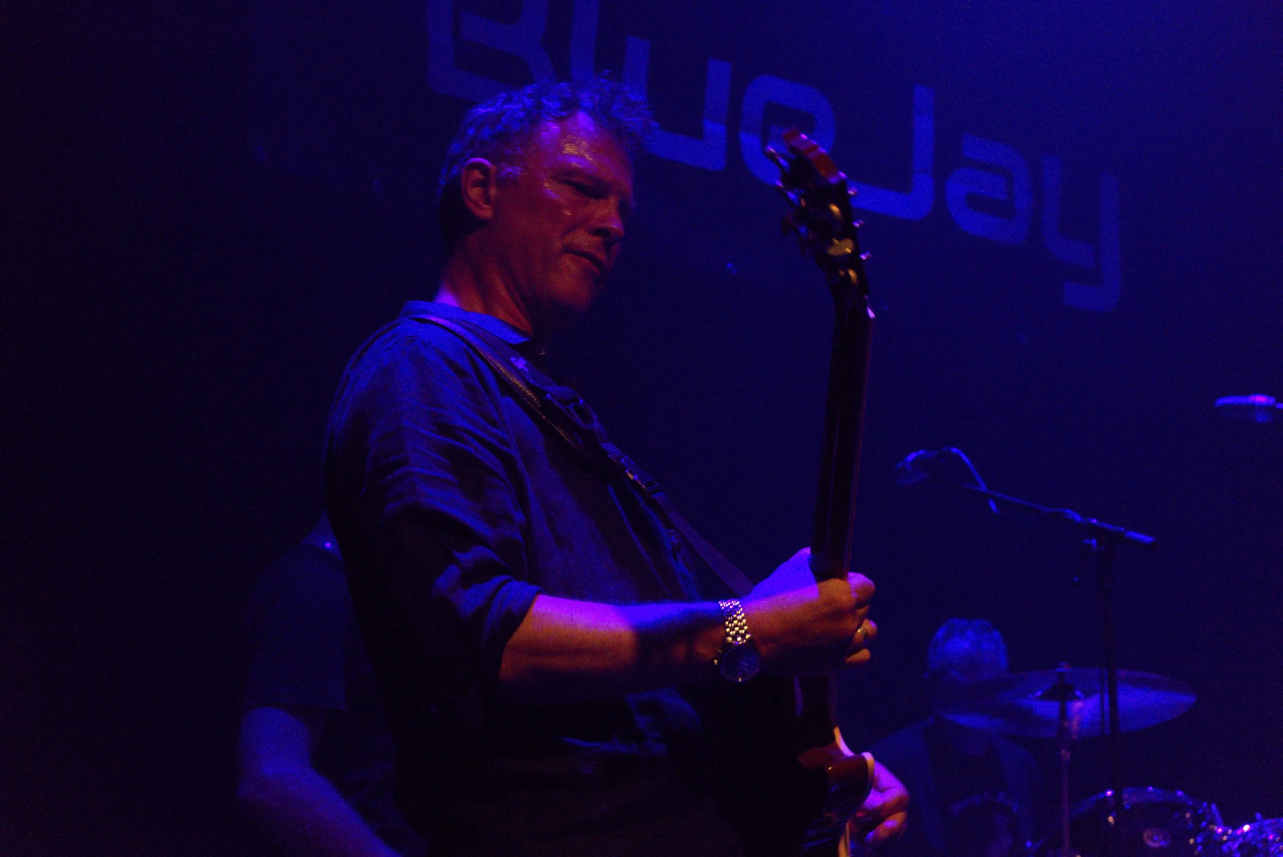 A man with gray hair playing an electric guitar on stage under blue lighting.