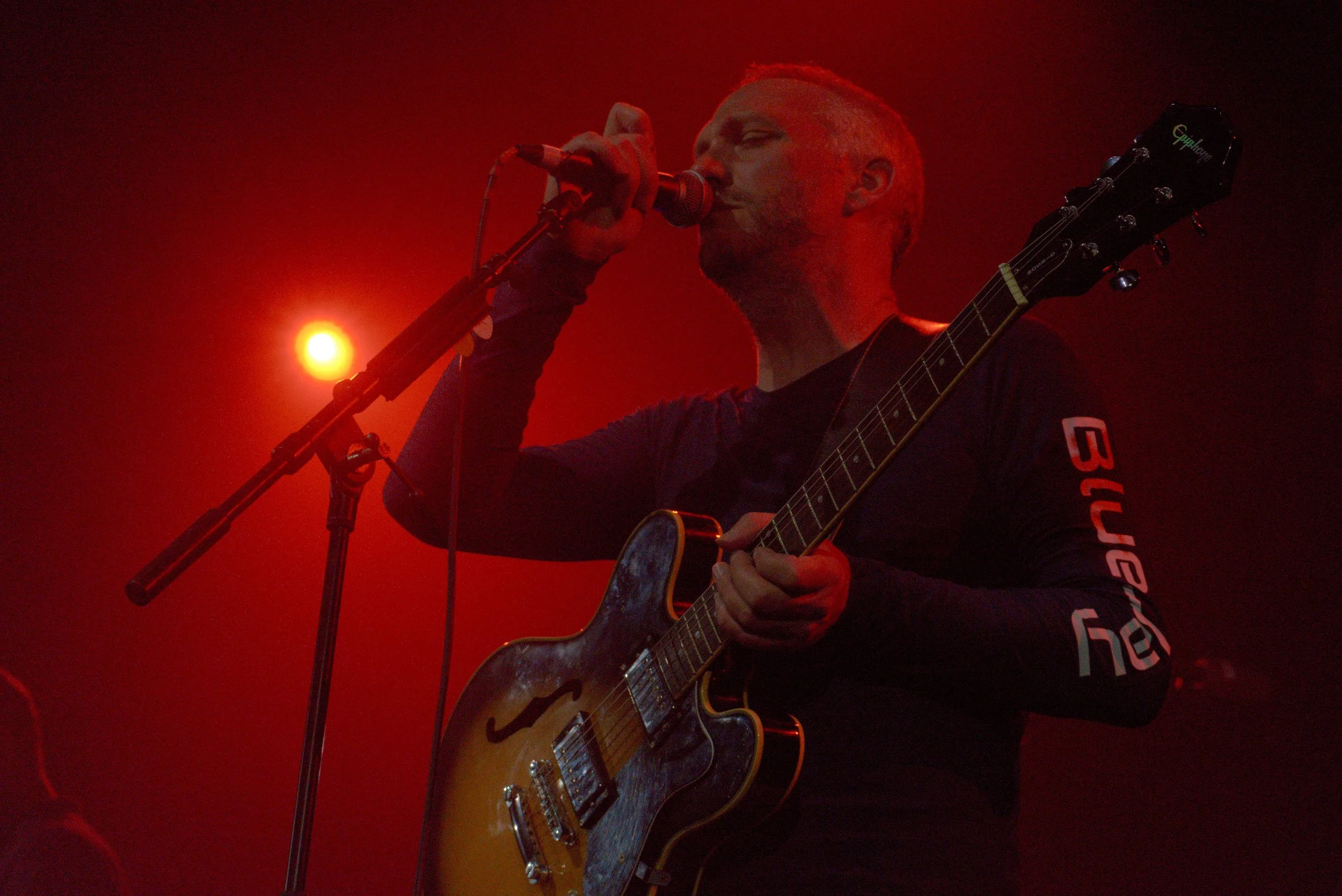 A male musician performing on stage, singing into a microphone and playing an electric guitar with a sunburst finish. The stage is lit with red lighting, creating a vibrant atmosphere.