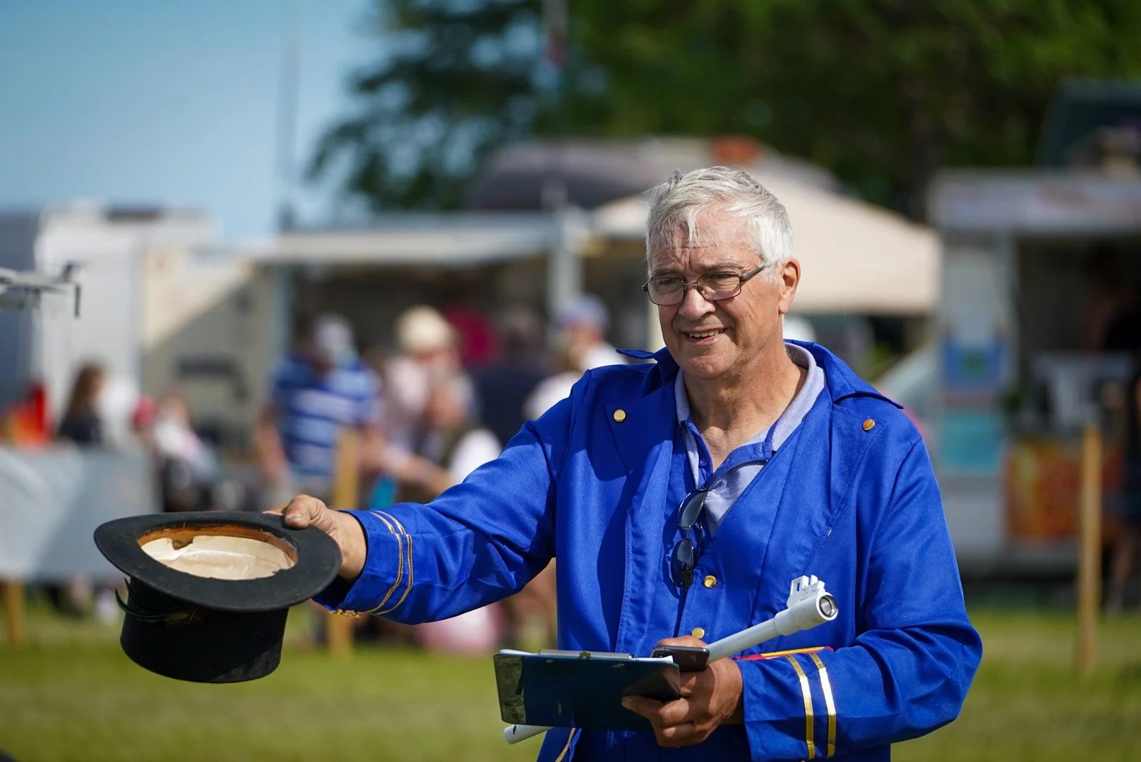 Older man with gray hair and glasses wearing a blue jacket with gold accents, holding a magician's hat and a magician's wand, at an outdoor event with tents and people in the background.