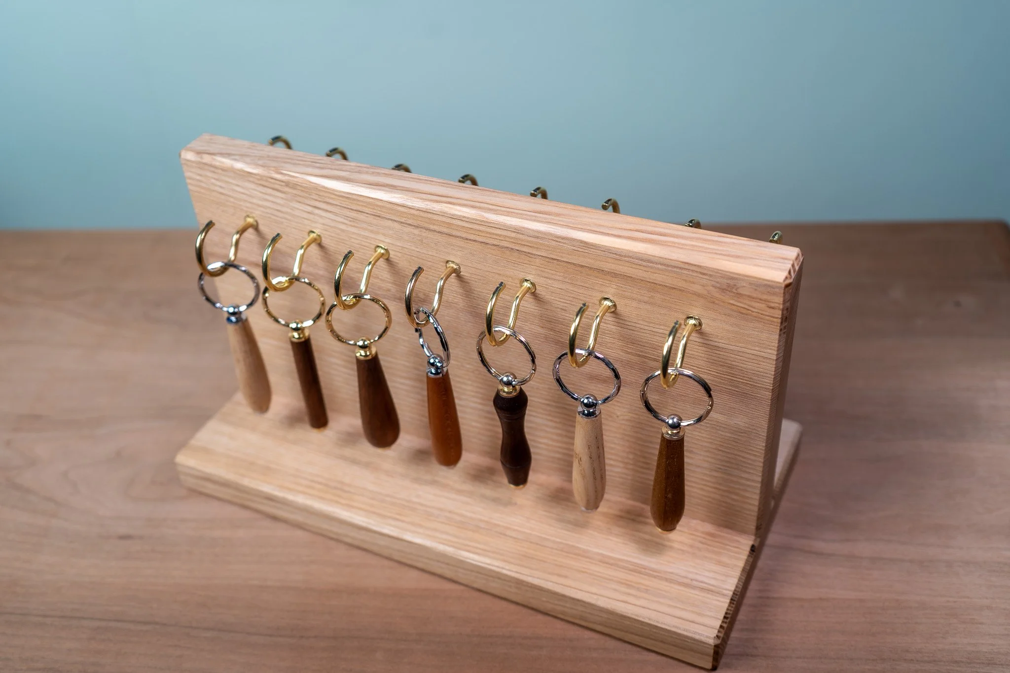 A wooden key holder stand with multiple hooks, holding several keys with wooden and metallic keychains, placed on a wooden surface with a plain blue wall in the background.