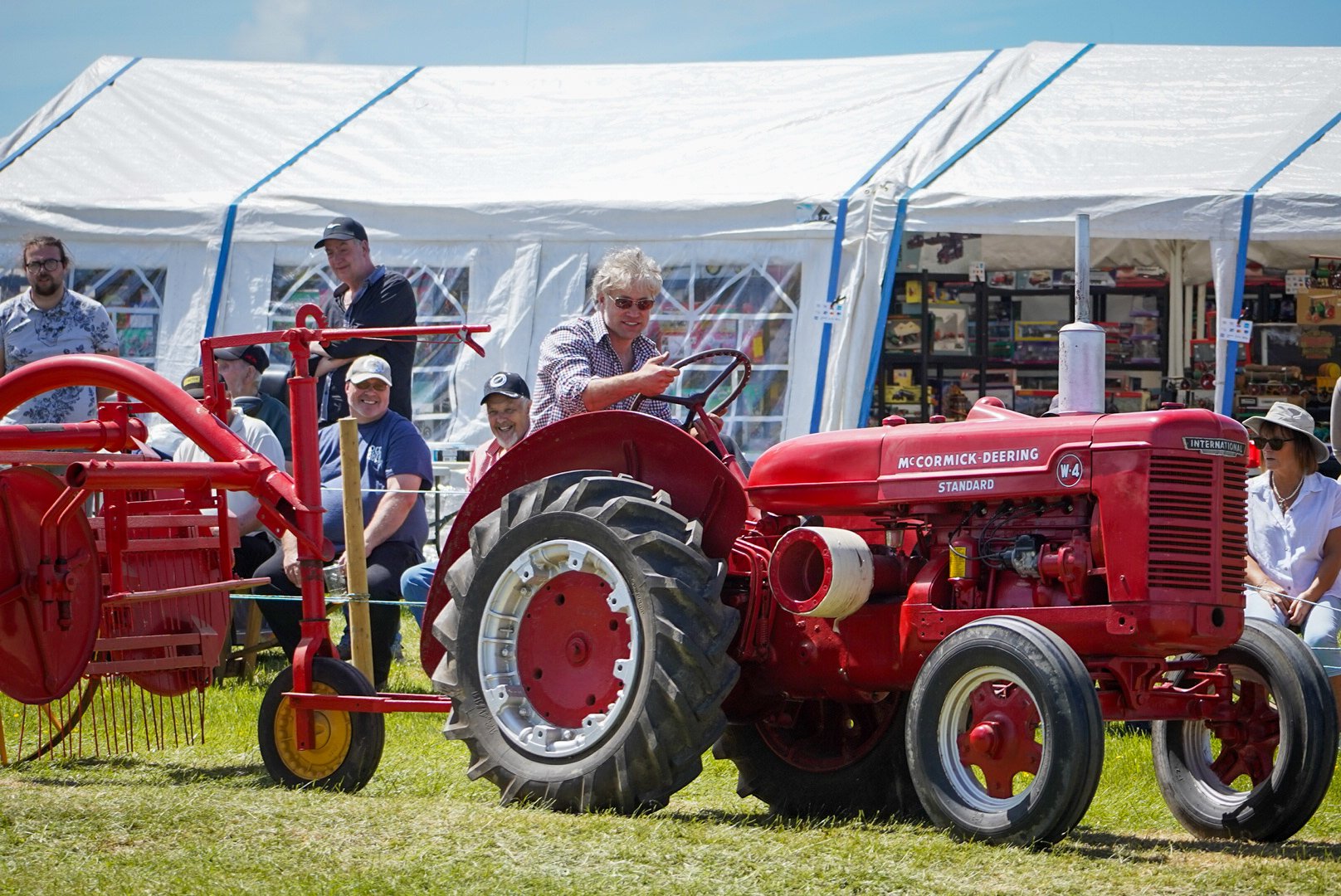 People sitting and standing near a vintage red McCormick-Deering tractor at an outdoor event, with a large white tent in the background.