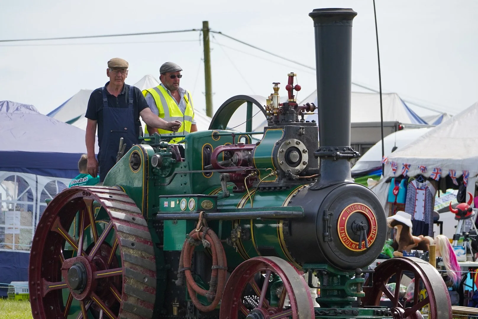 Two men examining a vintage steam engine at an outdoor event with tents and stalls in the background.