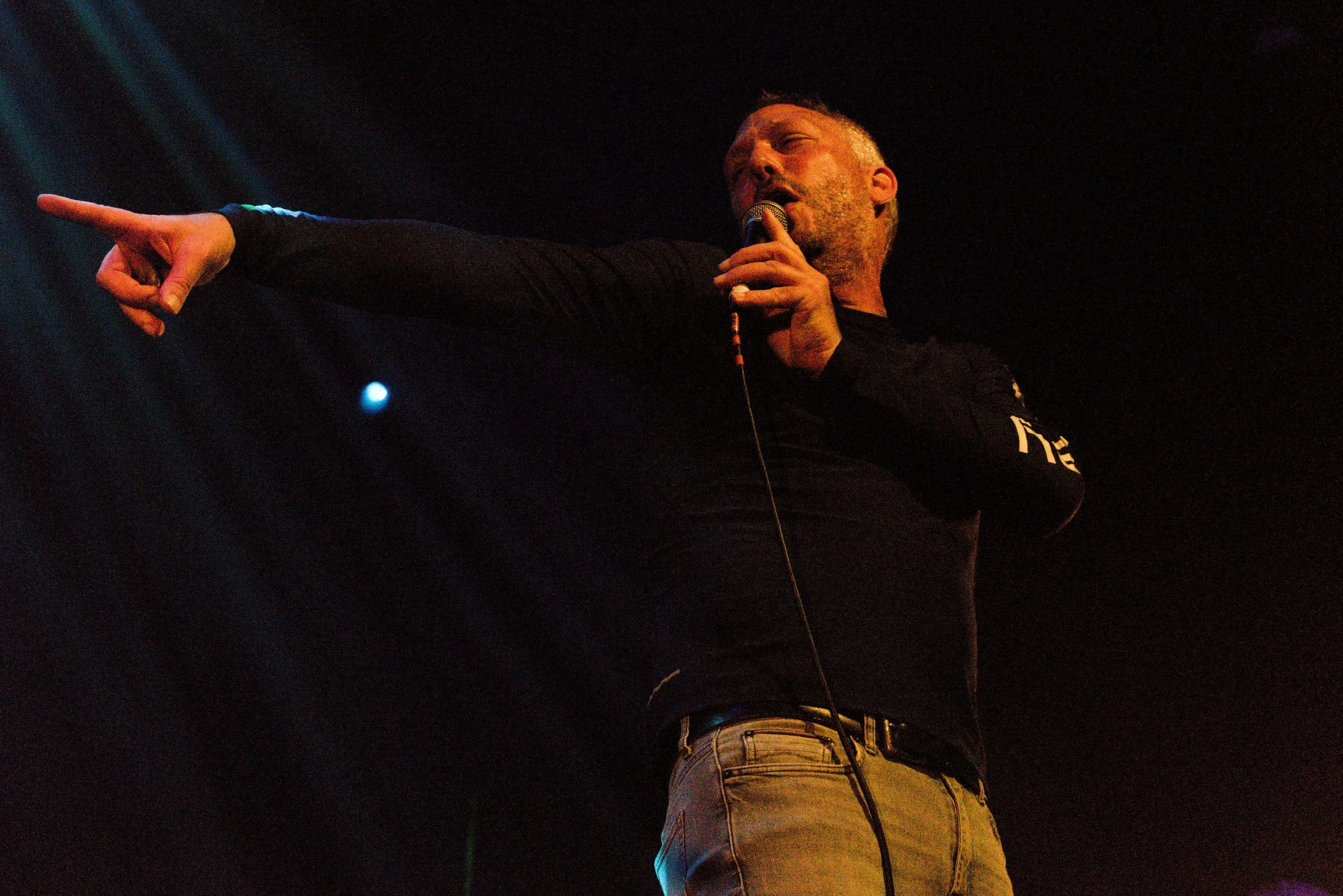 A man is performing on stage, holding a microphone in one hand, with his other arm extended and pointing. The background is dark with some stage lighting.