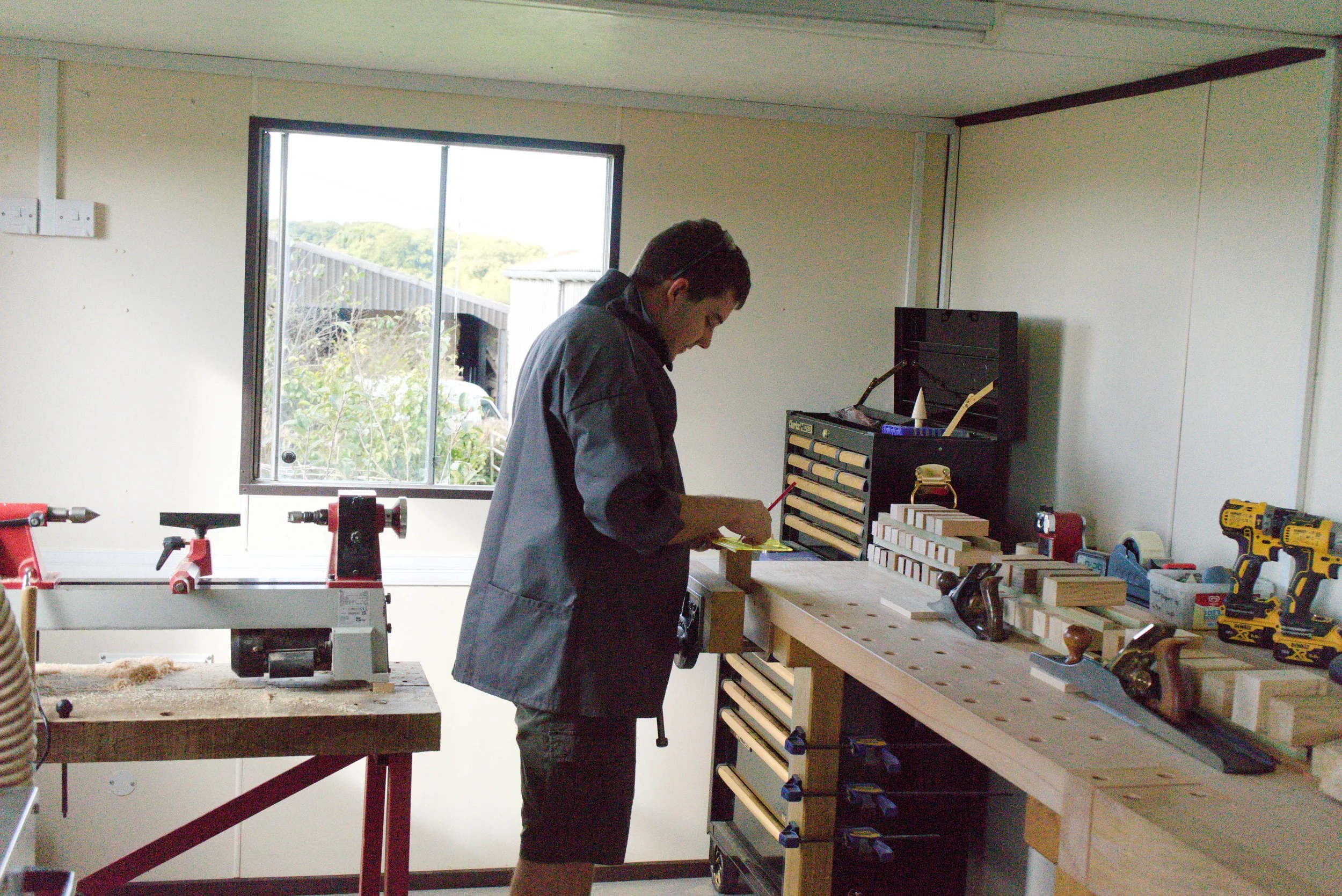 A man working in a woodworking shop, wearing a gray jacket, with various tools and wooden pieces on a workbench.