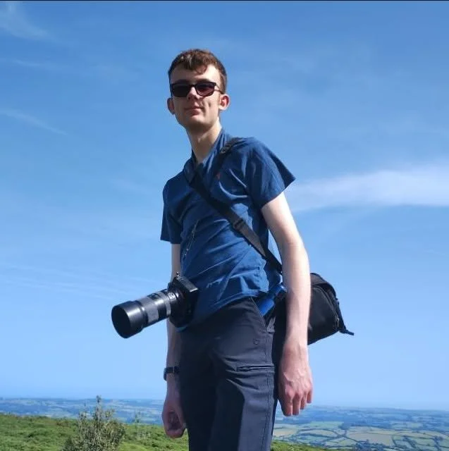 Young man with sunglasses, wearing a blue shirt and carrying a camera and backpack outdoors on a sunny day, with a landscape and blue sky in the background.