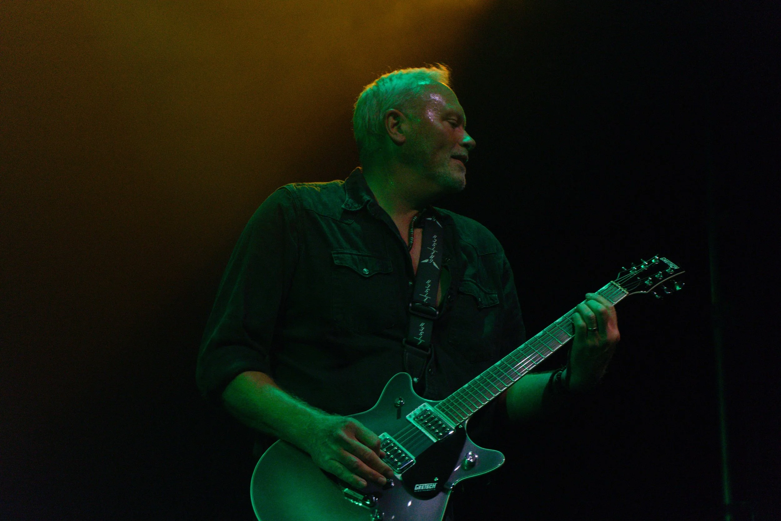 A man with silver hair and a beard playing an electric guitar on stage, illuminated by green and yellow stage lighting, with a dark background.