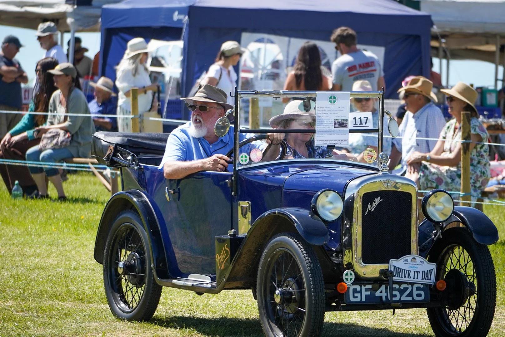 A vintage blue Austin car with a man and woman in hats, surrounded by people at an outdoor event on a sunny day.