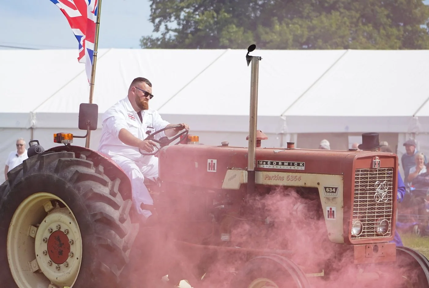 A man with sunglasses and a white outfit driving an old red tractor during an outdoor event, with people in the background and a Union Jack flag flying nearby.