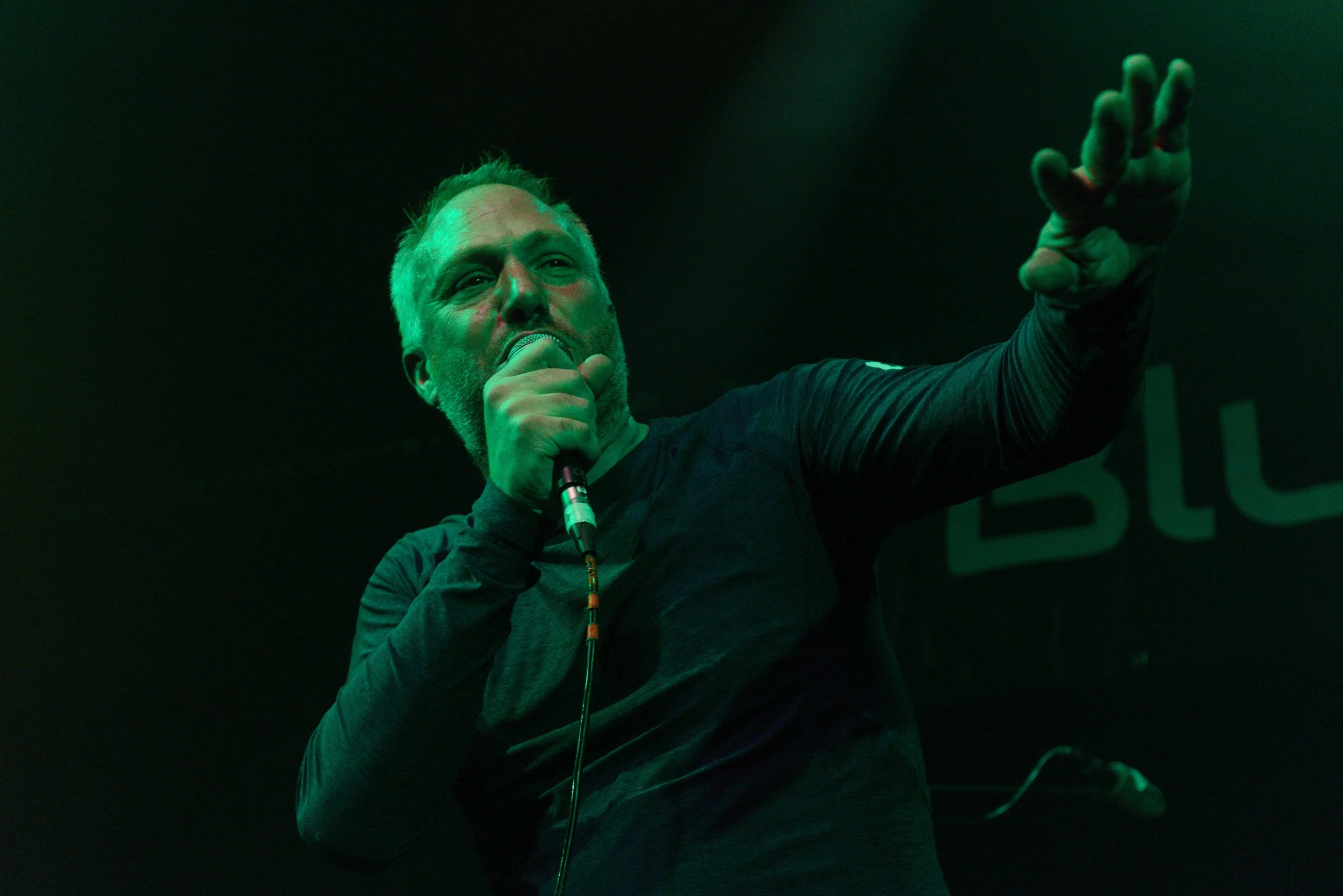 A man with gray hair and a beard is singing or speaking into a microphone on stage, with his arm raised and hand extended outwards. The scene is lit with green lighting and a dark background.