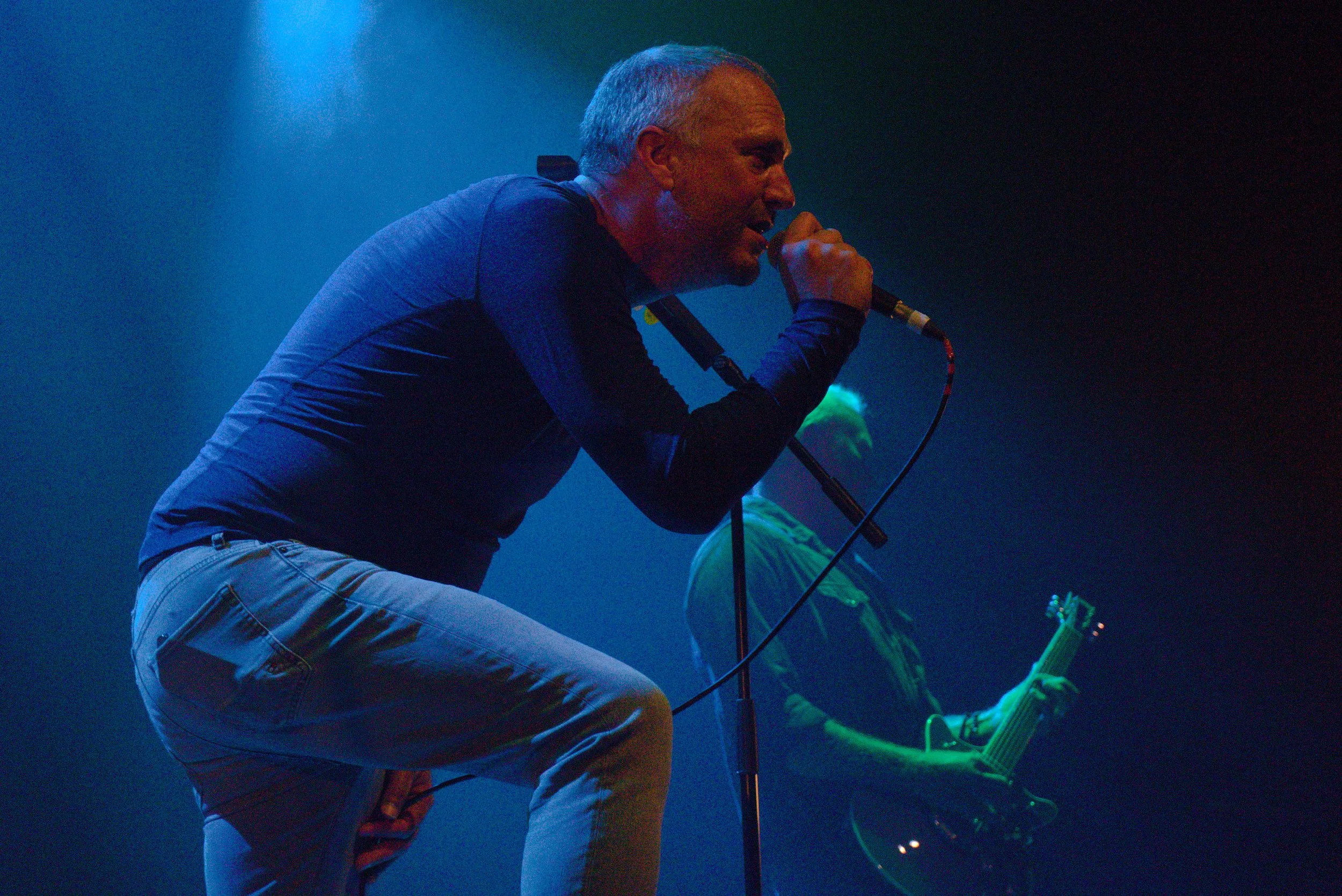A man with gray hair singing into a microphone on stage, with a guitarist in the background playing an electric guitar. The stage is dimly lit with blue and green lights.