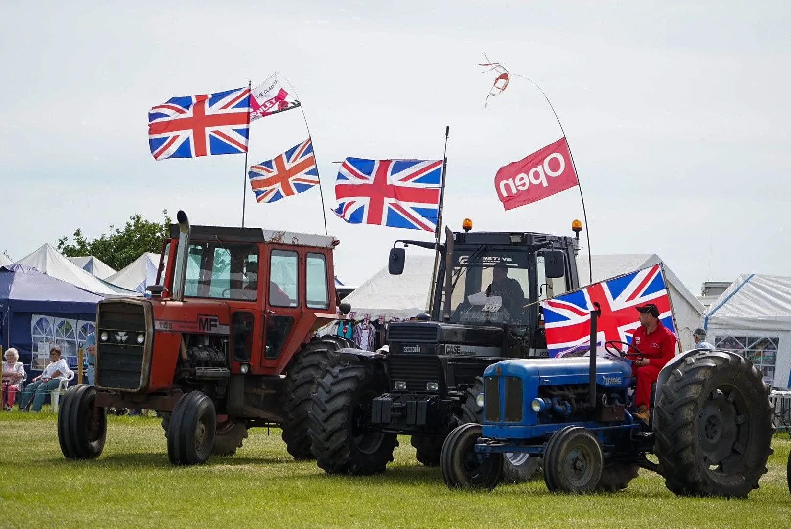 Two tractors, one red and one blue, decorated with Union Jack flags, are positioned side-by-side at an outdoor event. A person dressed in red sits on the blue tractor. Several tents and spectators are visible in the background.