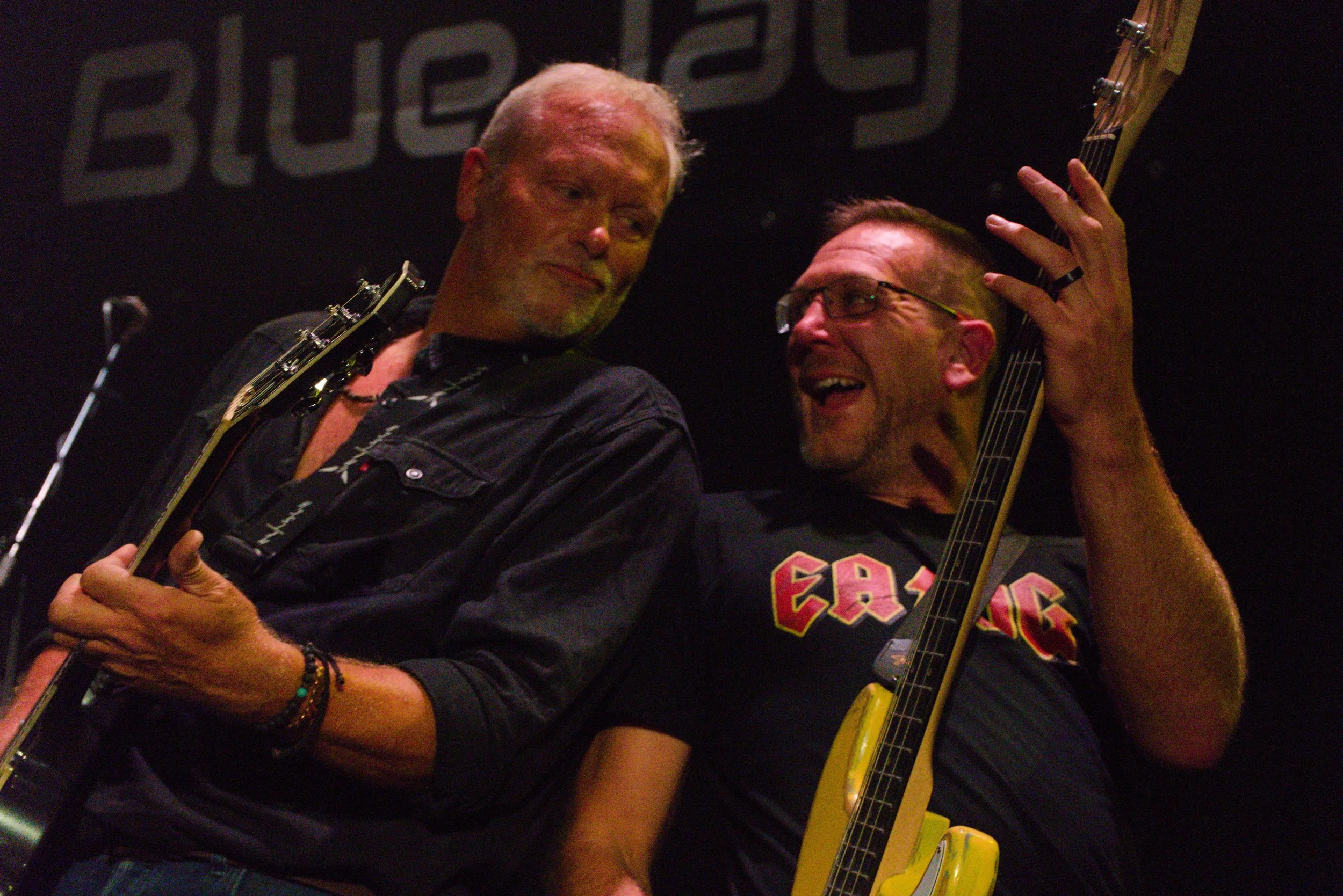 Two men playing electric guitars on stage, one with white hair and a grey beard, and the other with short dark hair and glasses, both smiling and engaging with each other.